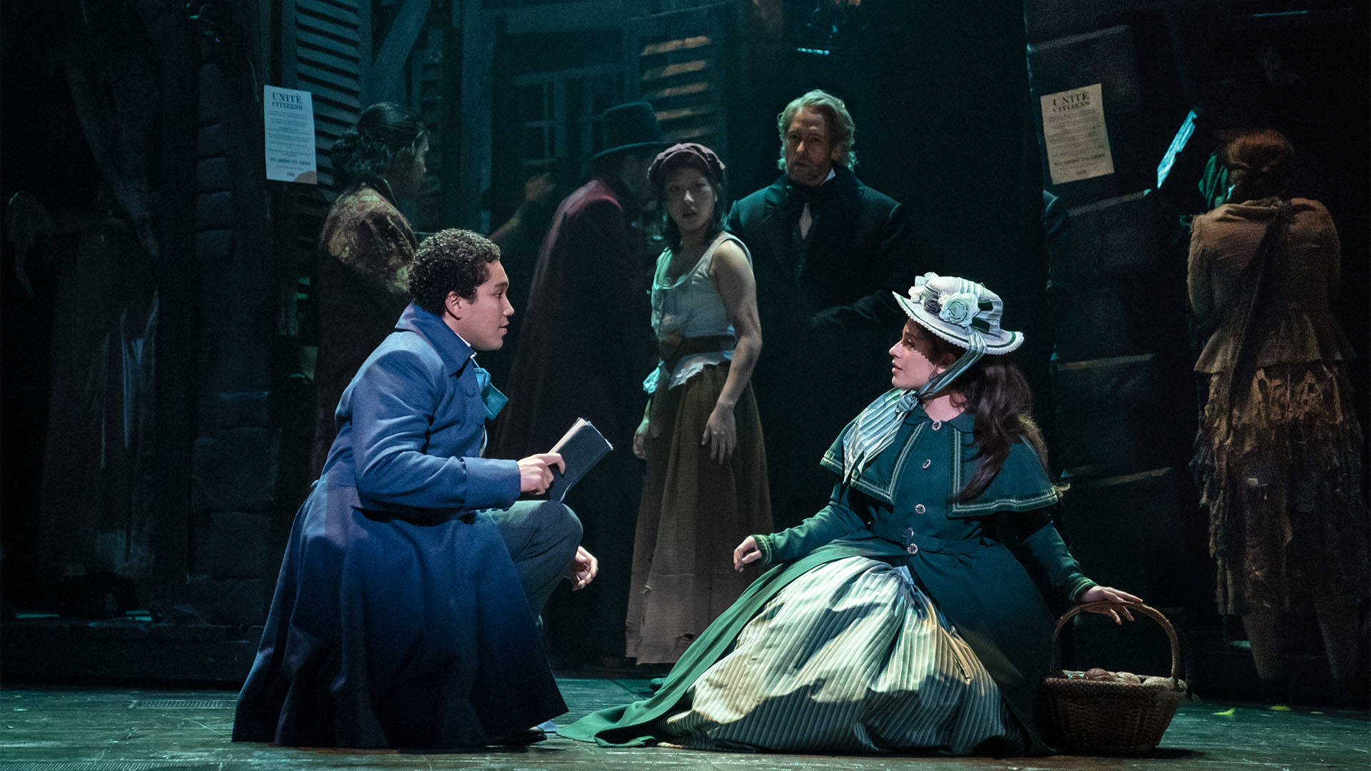 The cast of Les Miserables performing. At the center a man in a long blue coat holding a book squats and looks at a woman in a green dress.