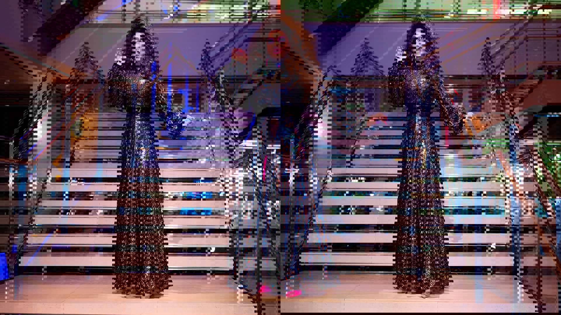 A drag performer in a dark outfit stands in front of a stone and glass staircase.