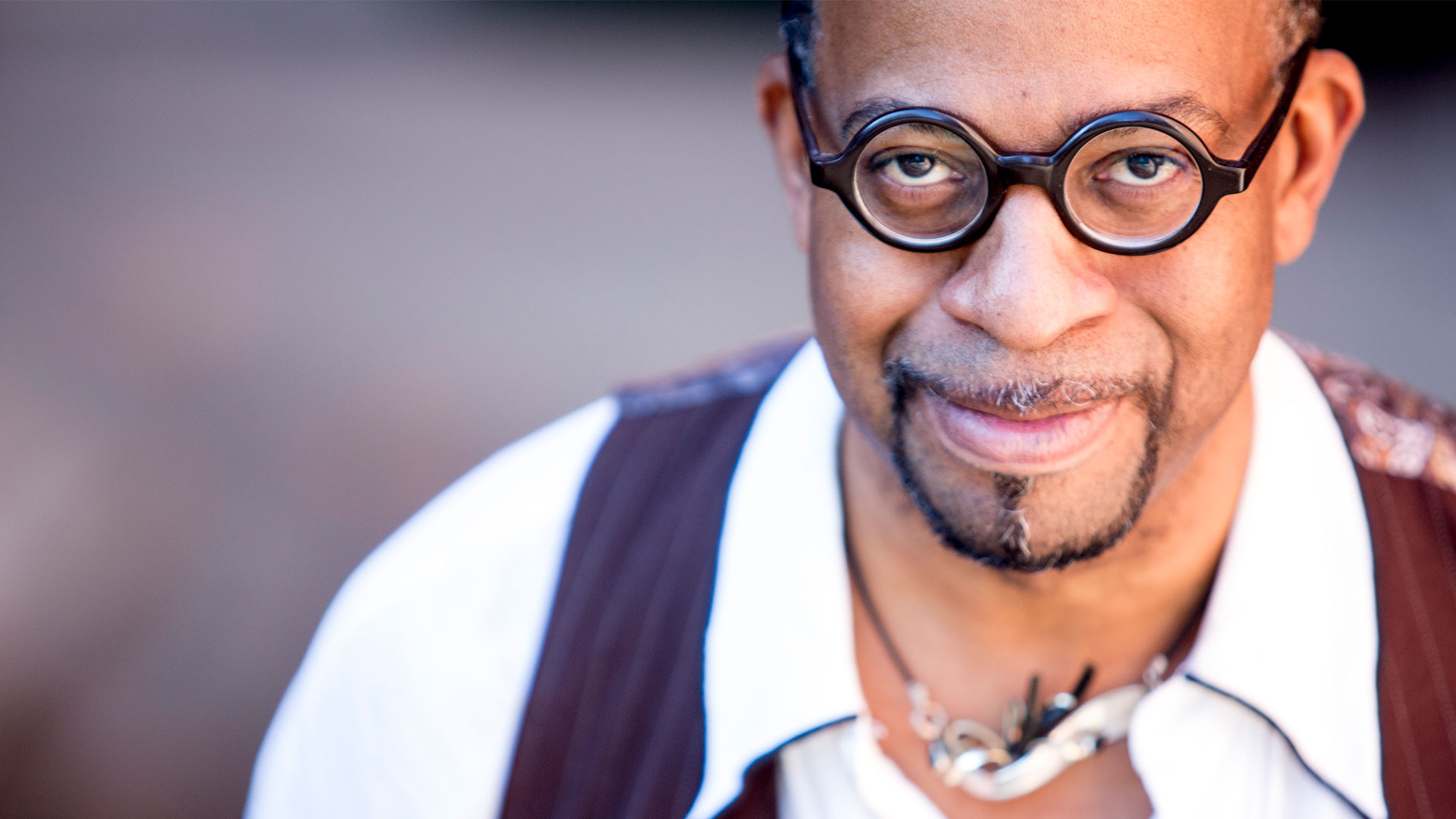 Headshot of a middleaged african american man with a goatee and glasses. He is wearing a collared white shirt with a burgundy vest as well as a necklace.
