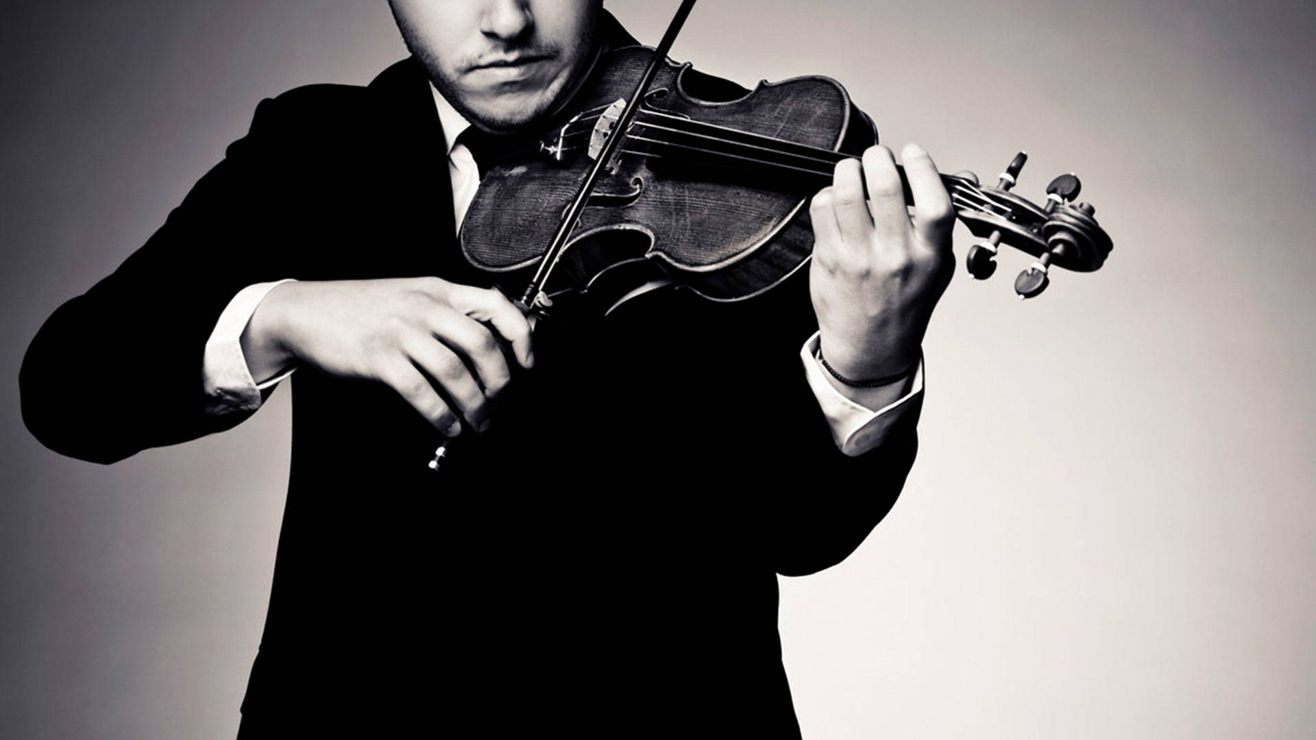 A greyscale photo of a man in a suit playing violin. He has light complexion and the photo is cropped such that only the lower half of his face is visible.