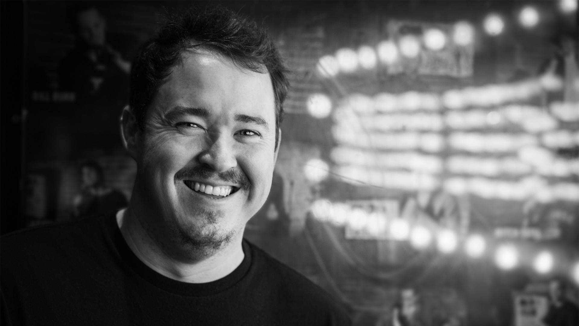 Grayscale headshot of a smiling man with light complexion, short hair and a light moustache and beard in a black shirt.
