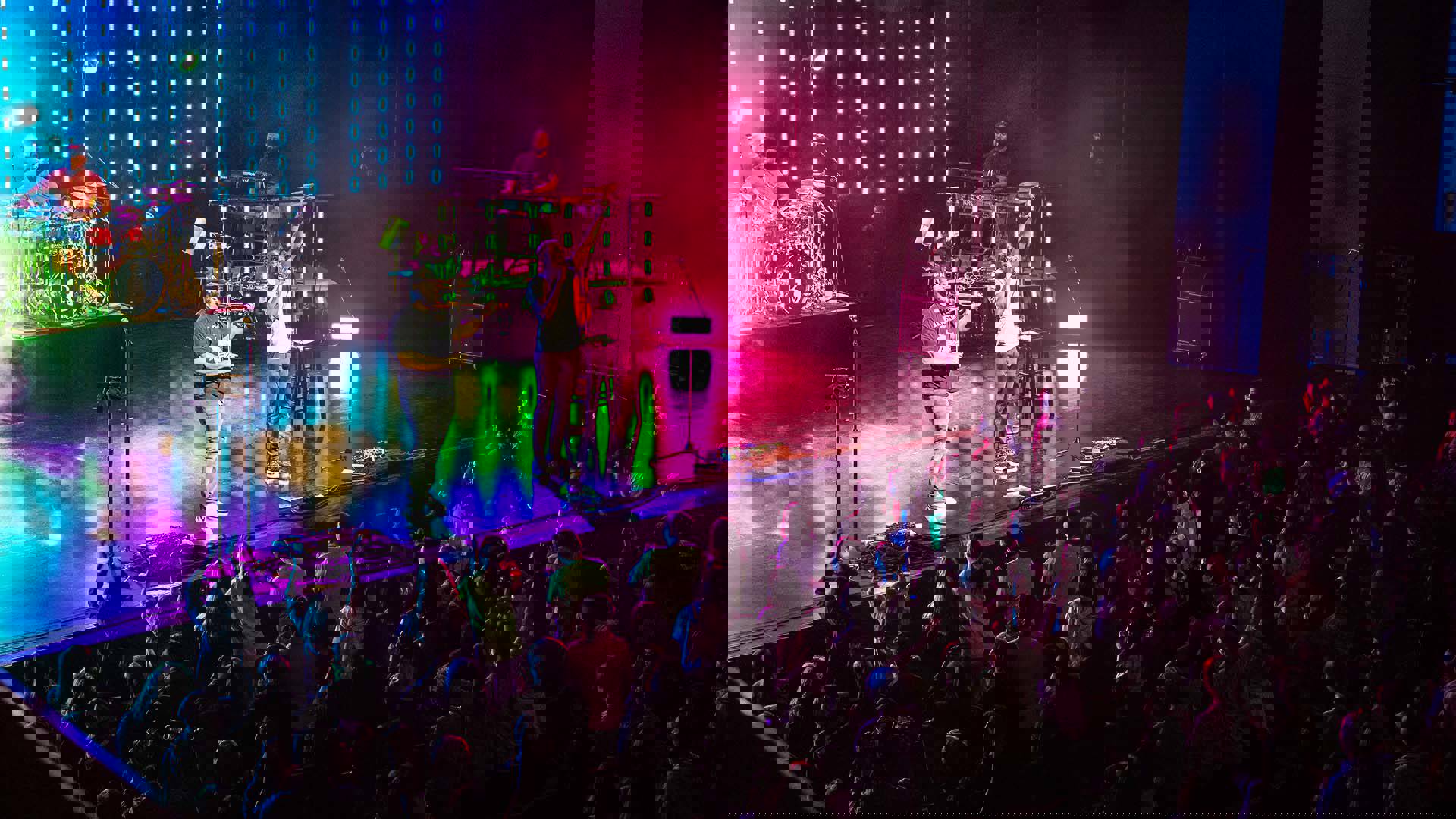 Black Violin performing on a colorful stage. Two lead perfomers are at the front of the stage. On the left, a black man playing a violin with a beard, black hat, and black shirt. On the right a black man speaking into a microphone with a white shirt on and holding his hand in the air., The young audience is on their feet and excited.
