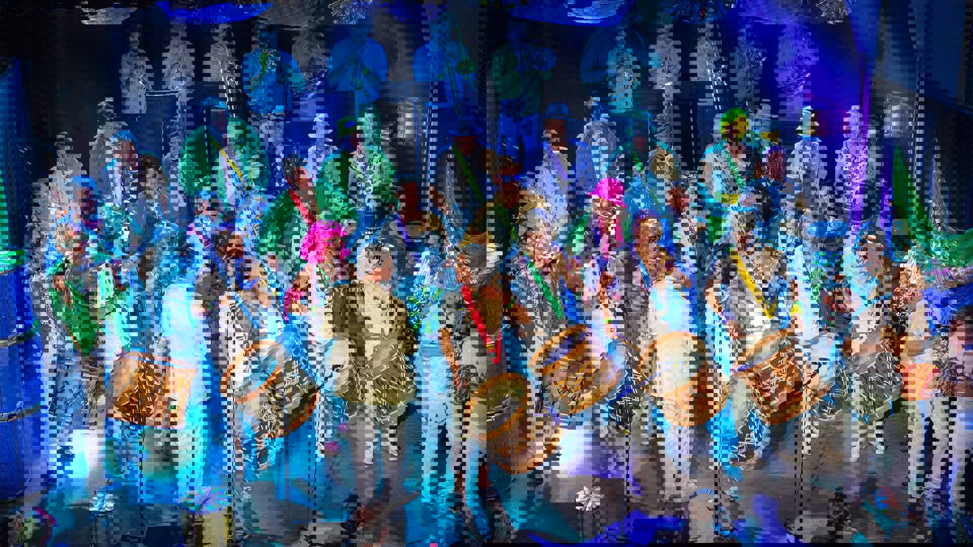 A large group of percussion and brass musicians wearing white and green performing on a blue lit stage.