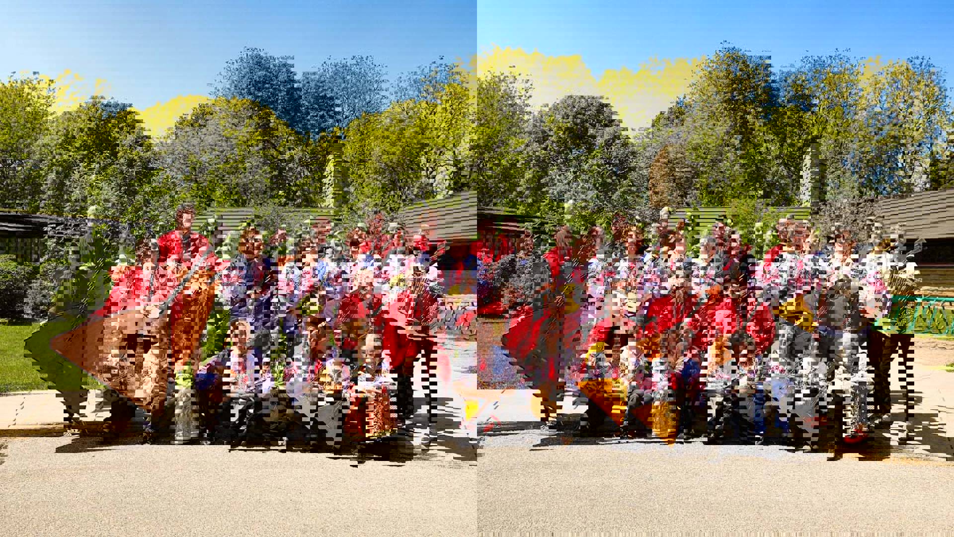 Several dozen russian folk orchestra musicians in red and white outfits pose outside for a picture. They are holding string instruments.