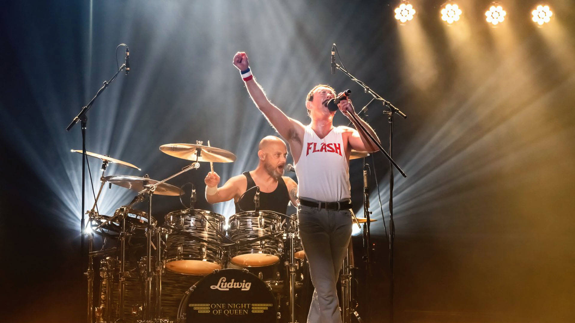 A Freddie Mercury impersonator wearing a white tank top with the word "FLASH". He has light complexion and short brown hair. He is singing into a microphone with his fist in the air. There is a drummer behind him.