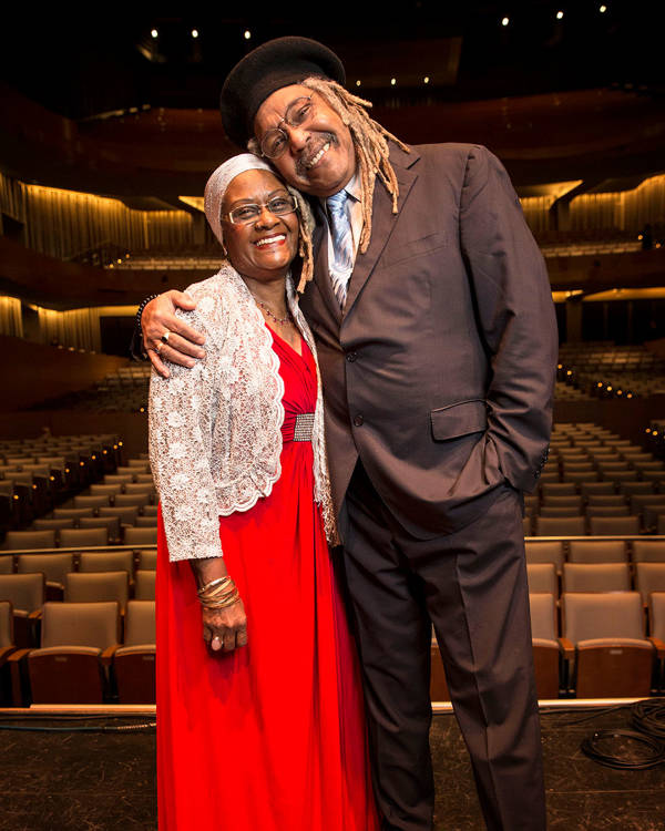 A man and a woman in the Afro Cuban Allstars embrace warmly and smile for a photo. The man is wearing a suit and the woman is wearing a red dress.