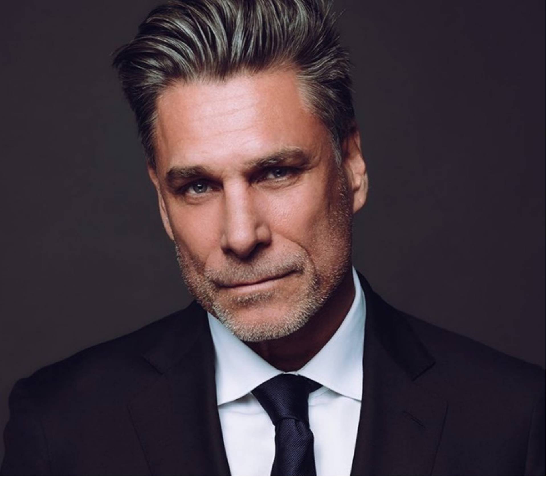 Headshot of a middle aged caucasian man with styled greyish black hair, a light grey beard and dark eyes. He is wearing a black suit and tie.