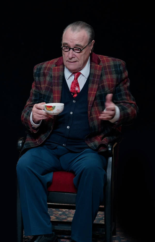 An older, white man with a receding hairline and black glasses in a blue suit and plaid jacket. He is sitting and holding a teacup.
