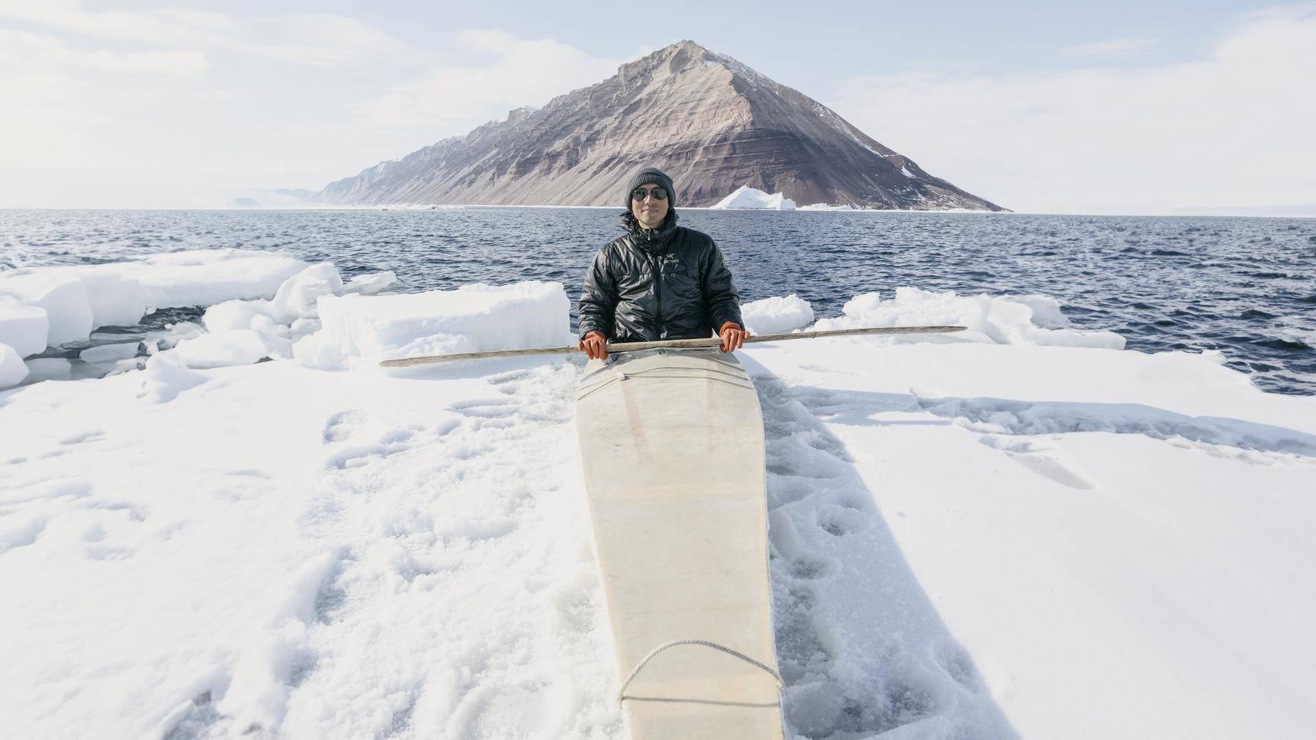 Photographer Kiliii Yuyan sits in a long white kayak on an ice berg. He is wearing dark glasses, a black hat and coat and orange gloves.