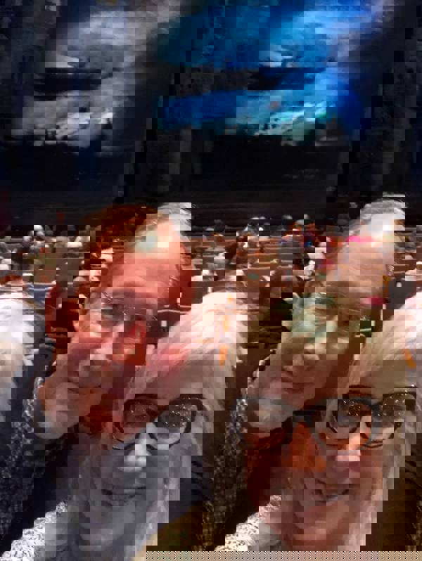 Selfie of a man and a woman with light complexion and grey hair in front of seats and a dark stage.