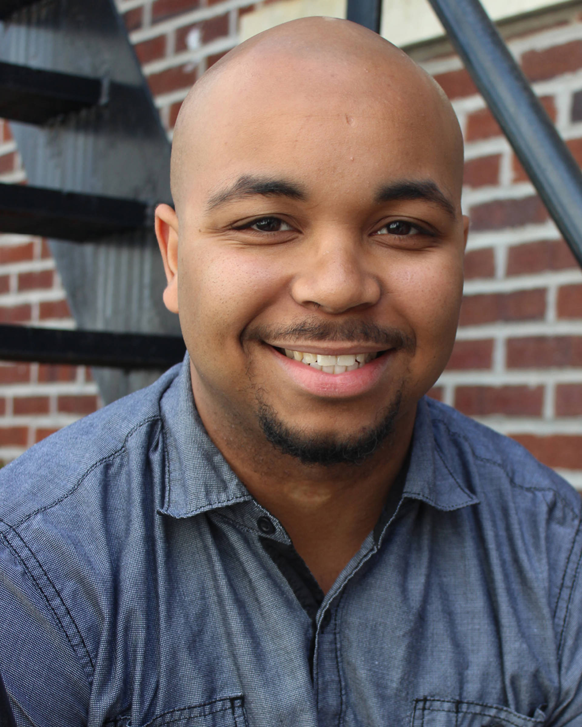 Headshot of an African American man with a bald head and brown eyes. He is wearing a collared blue shirt.