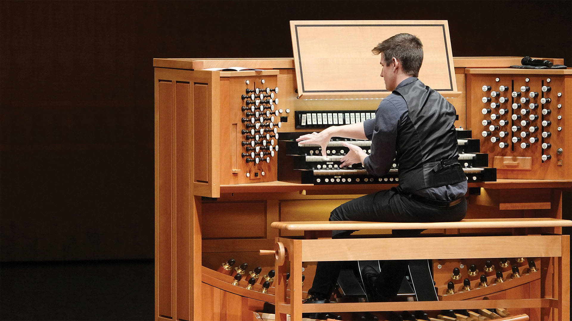 Greg Zelek, a young man with light complexion, short styled brown hair, a black button-up shirt and black vest, playing a large wooden organ console.