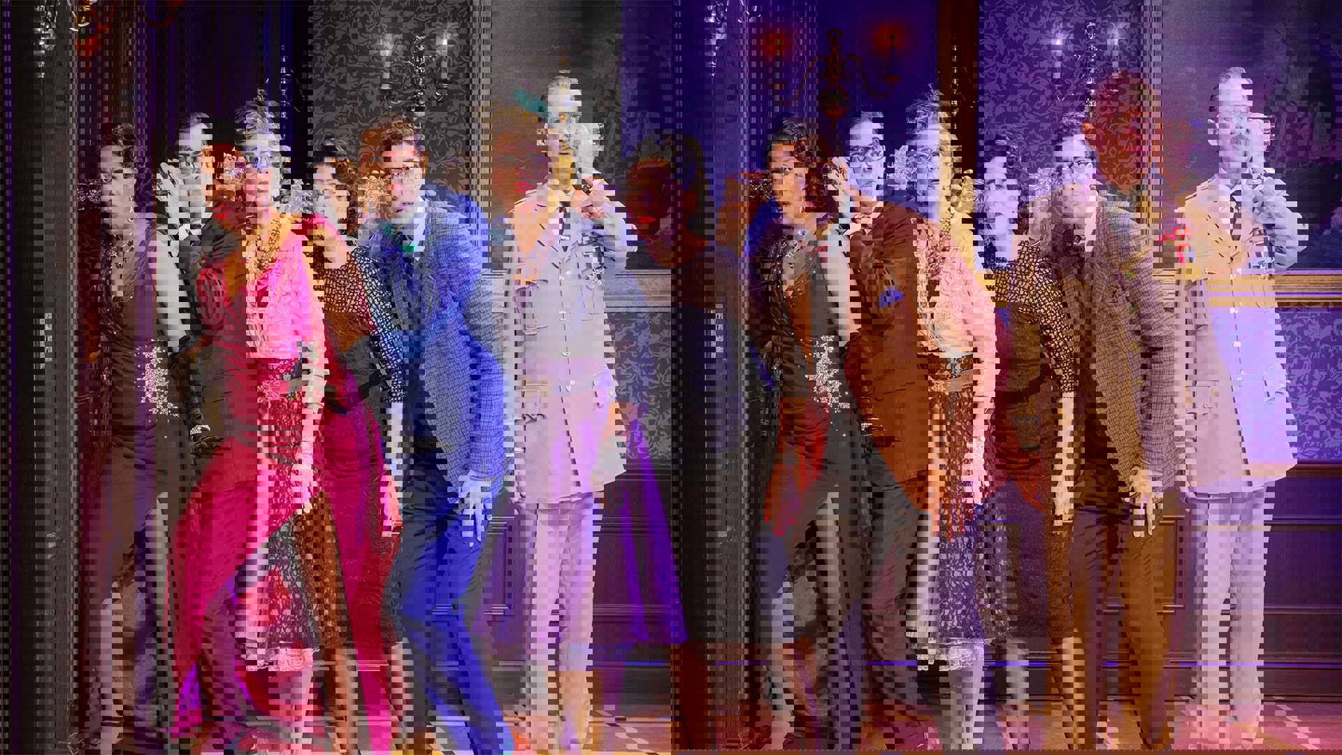 Six actors stand on stage in a line on one side of a closed wooden door, five of them are crouched close together with their ears pressed against glasses, trying to hear a conversation inside the closed door. The other actor is standing facing away from the rest, his glass pressed to his own ear, but the other end is connected to nothing.