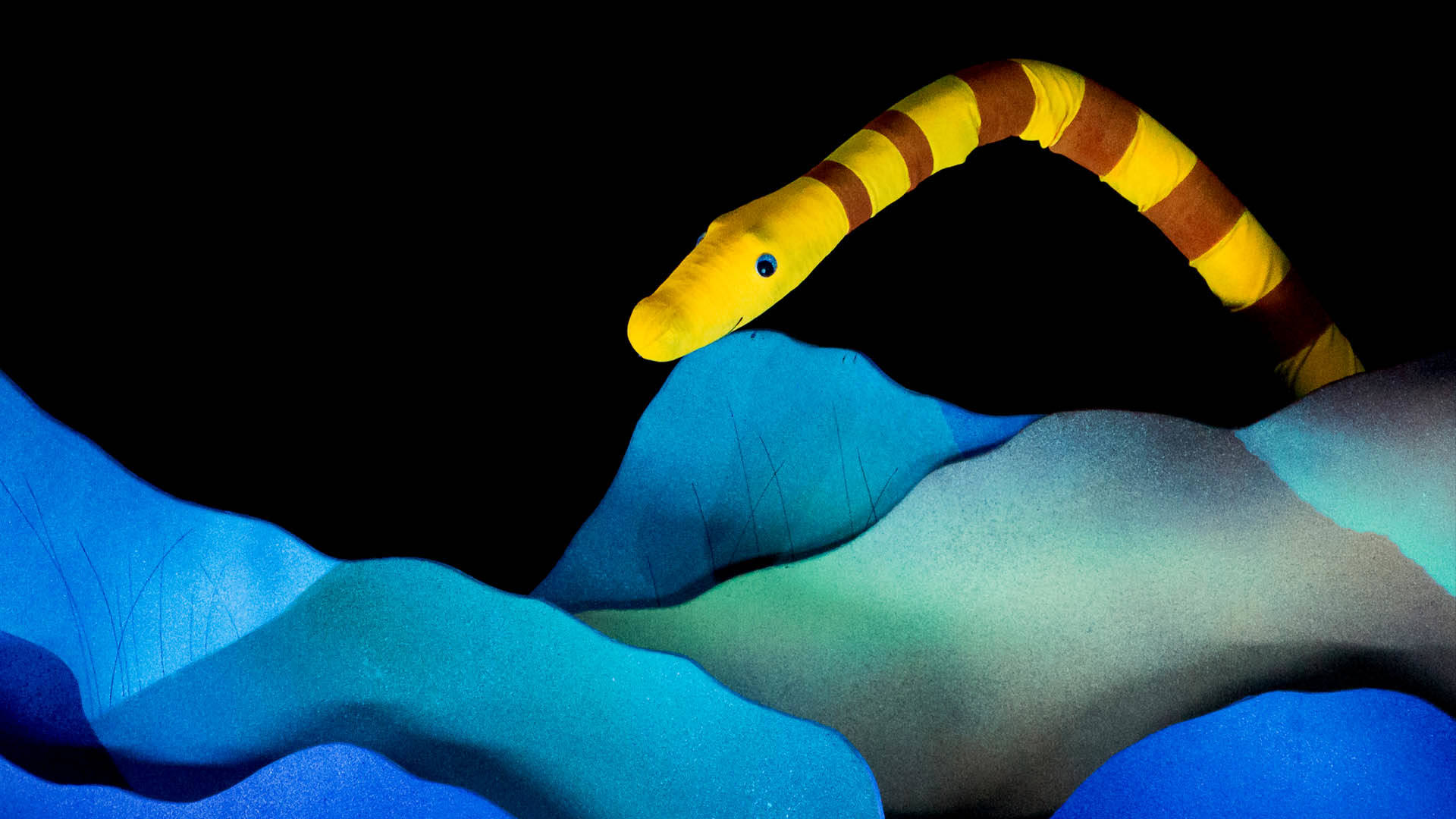 A puppet of a yellow and brown striped eel. It is sneaking out from behind blue bedrock.