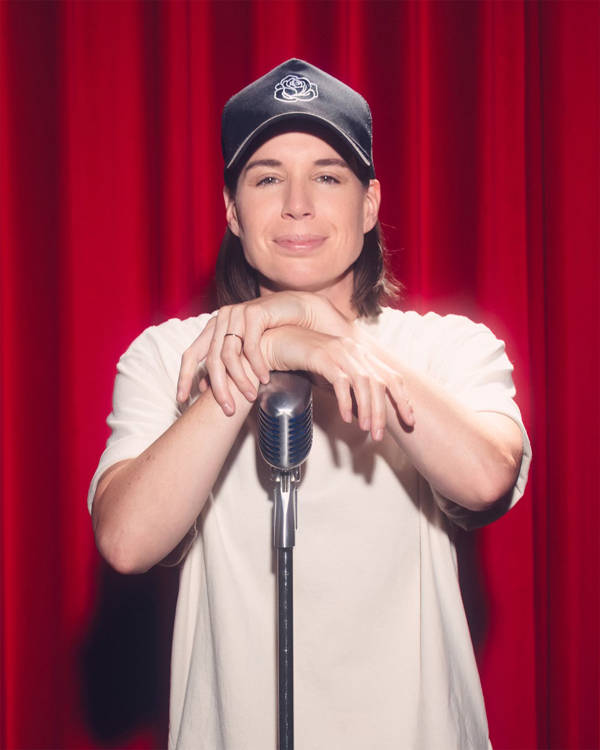 Danae Hays, a woman with light complexion, medium length brown hair, a baseball cap and a white tshirt, smiling and resting her arms on a microphone on a stand.