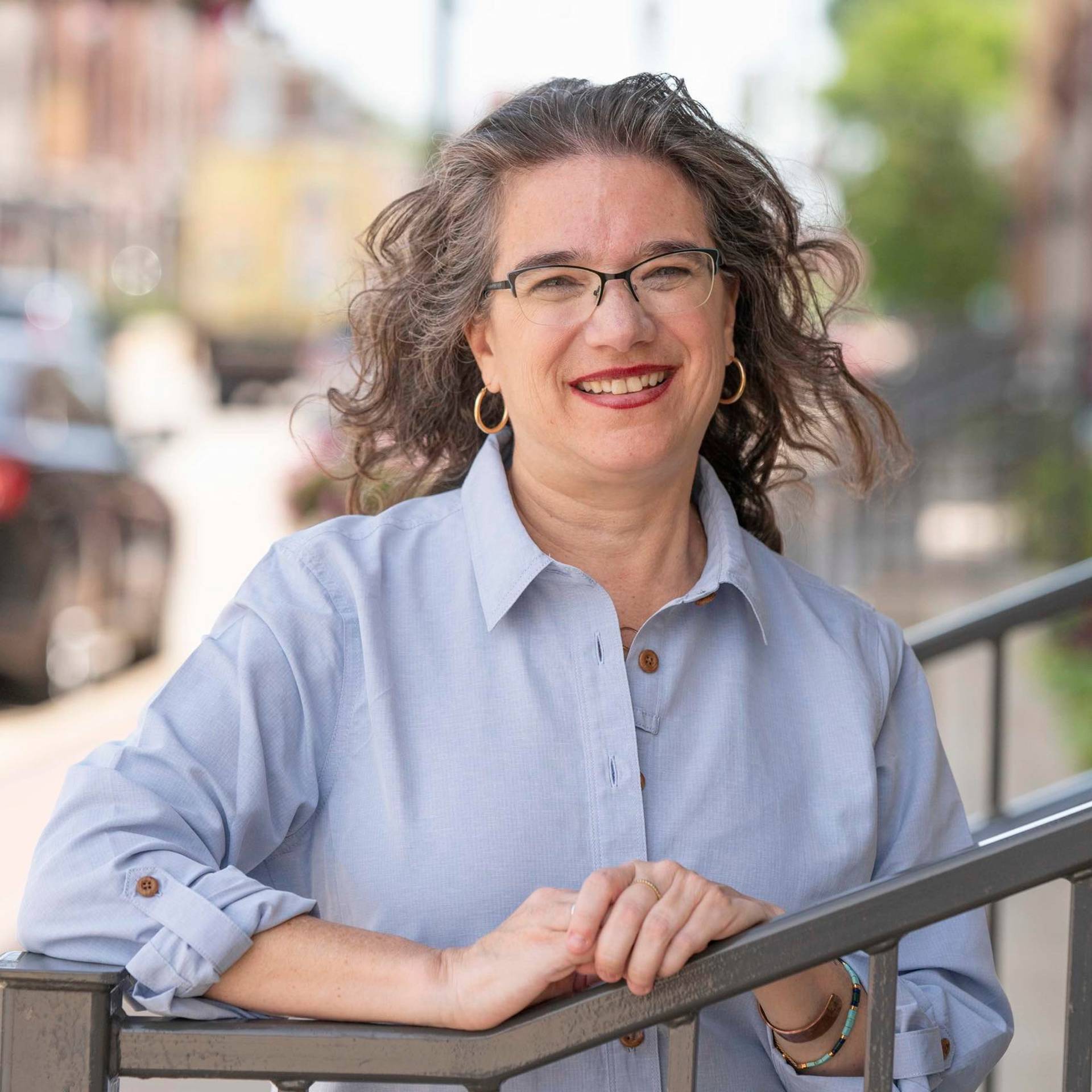 Melissa Agard Headshot. She is a middle aged woman with light complexion, wavy greyish brown hair, glasses, and red lipstick. She is wearing a pale blue button up shirt.