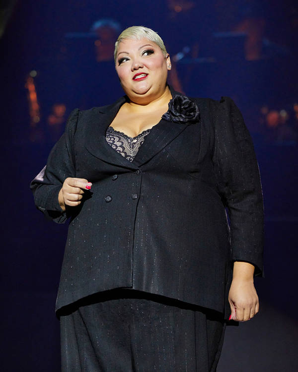 A woman in a pinstriped black pantsuit with short bleached hair stands on stage.