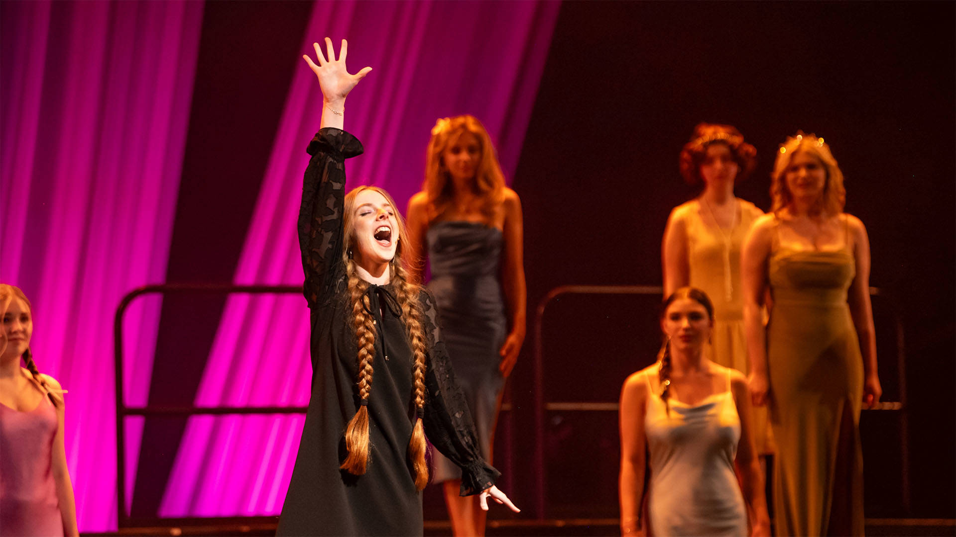 A student with light complexion, two long light brown braids in a black long-sleeved dress, singing with one hand raised and eyes closed.