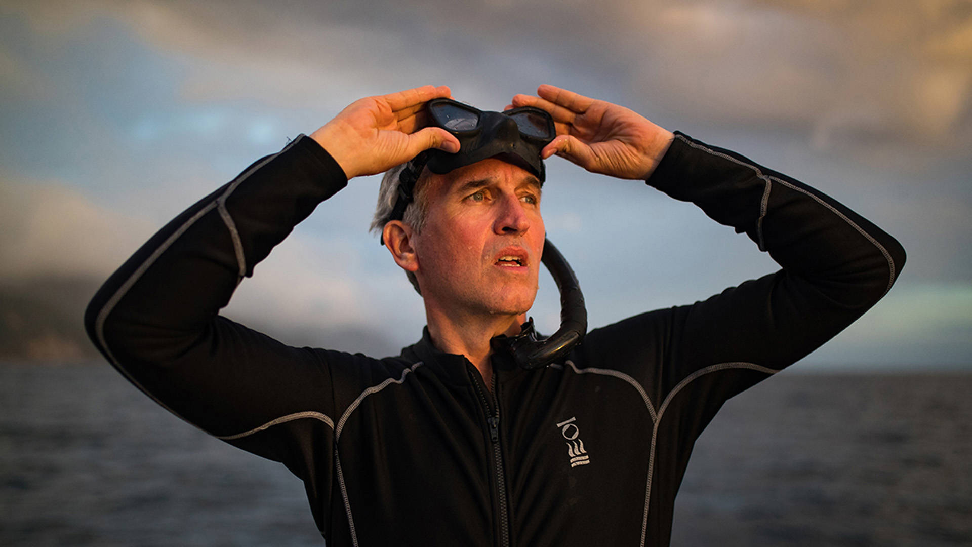 Brian Skerry, a caucasian man with short grey hair, wearing a black diving suit while pulling off snorkel goggles to his forehead with both hands.