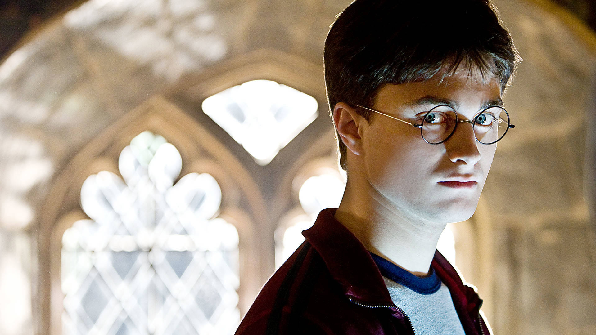 Harry Potter, a caucasian young man with short brown hair and circular glasses. He is looking at the camera and wearing a grey shirt and maroon athletic jacket.