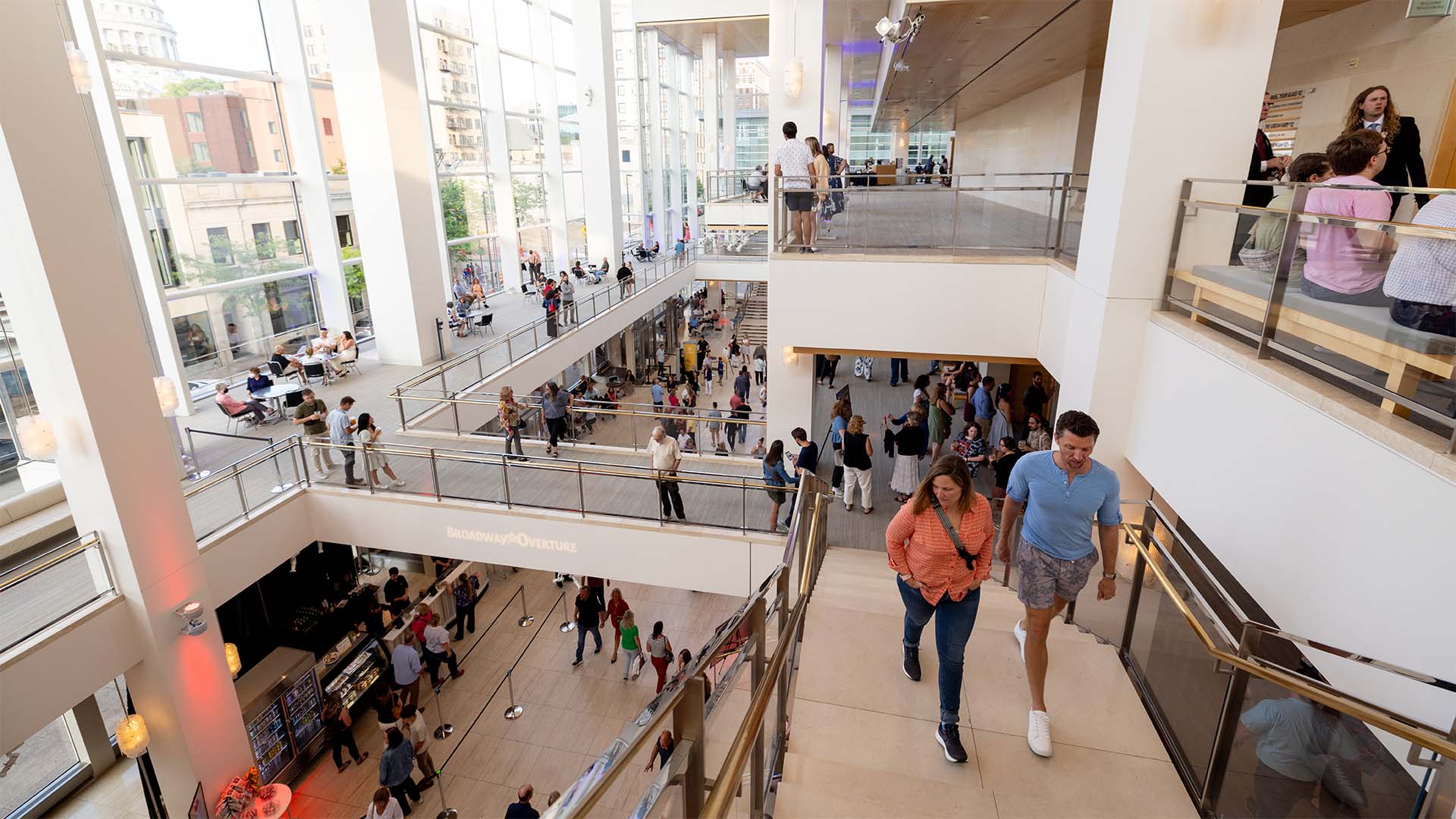 High view of Overture Hall Main Lobby, a large room with a rightside wall of windows facing Madison's downtown. People are crowded on the floor below in lines for concessions.