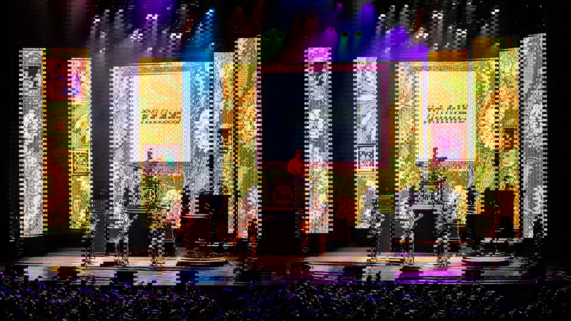 Four Beatles impersonators performing in front of a full house. Three are seated at the front of the stage holding guitars. A flowery wallpaper design is projected behind them.