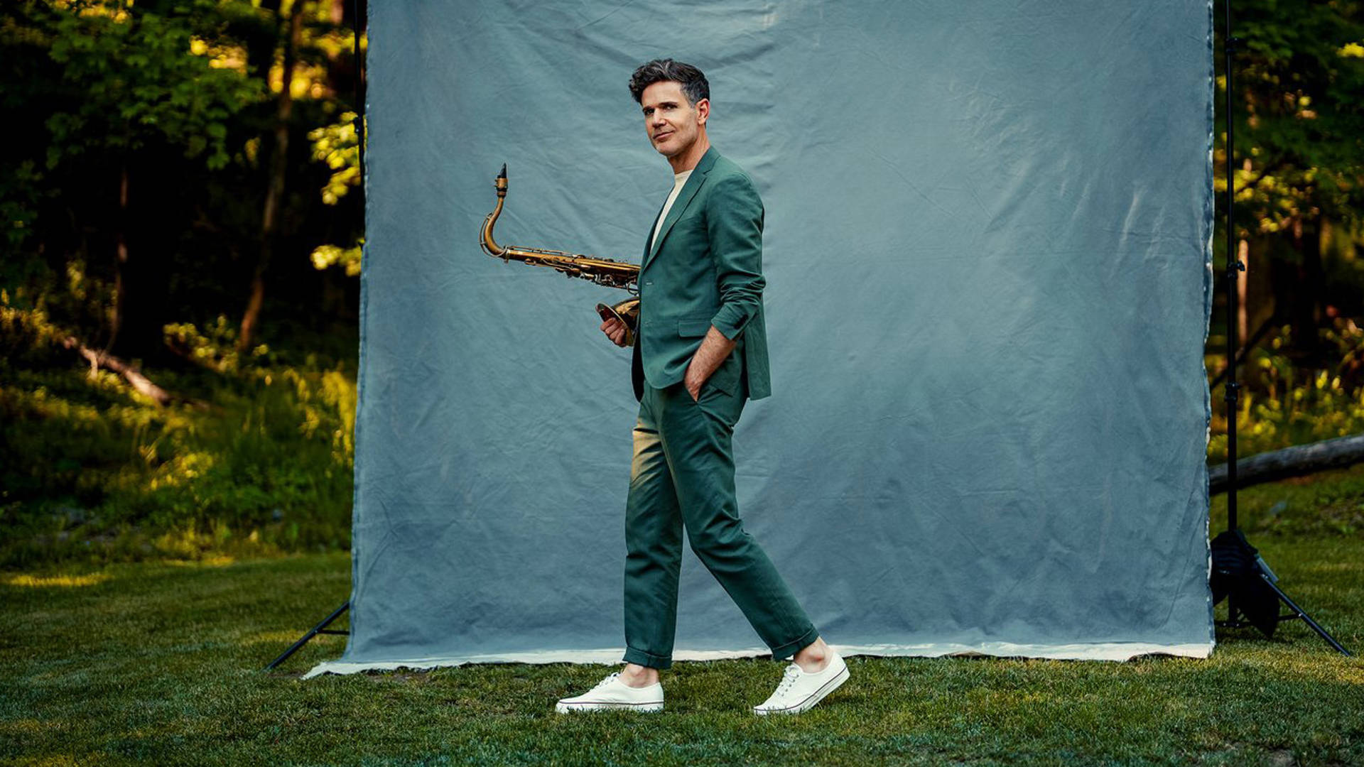 A young man with light complexion, short dark hair, and a teal suit walking on grass with a blue photography background set up behind him.