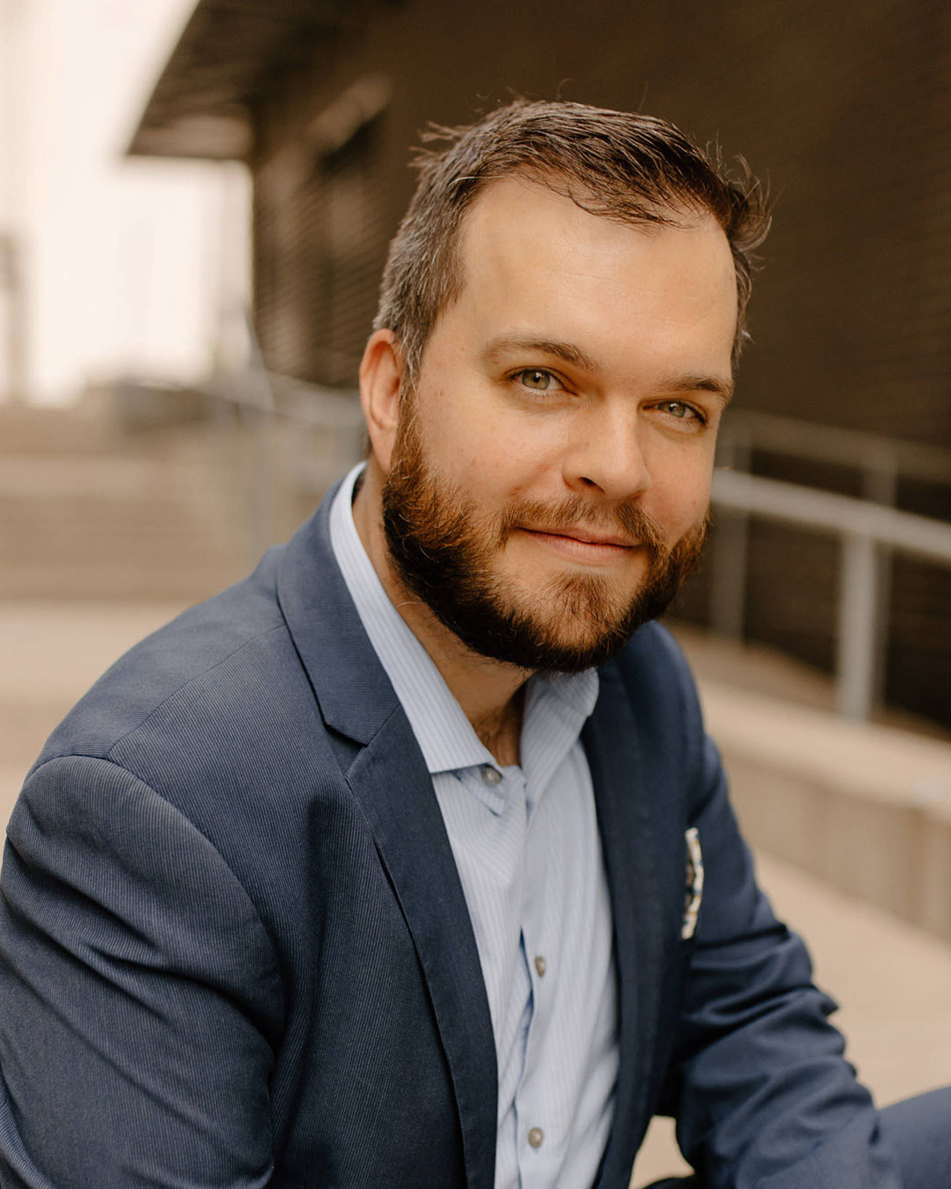 Headshot of a man with light complexion, short brown hair , hazel eyes, and a greyish blue suit.