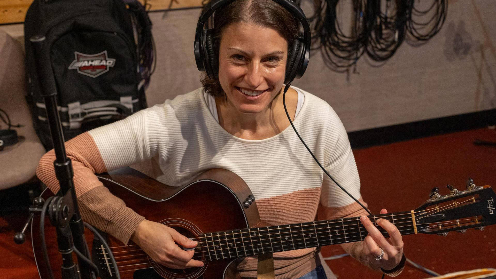 A woman with light complexion and brown hair seated playing an acoustic guitar. She has headphones on and is smiling toward the viewer.