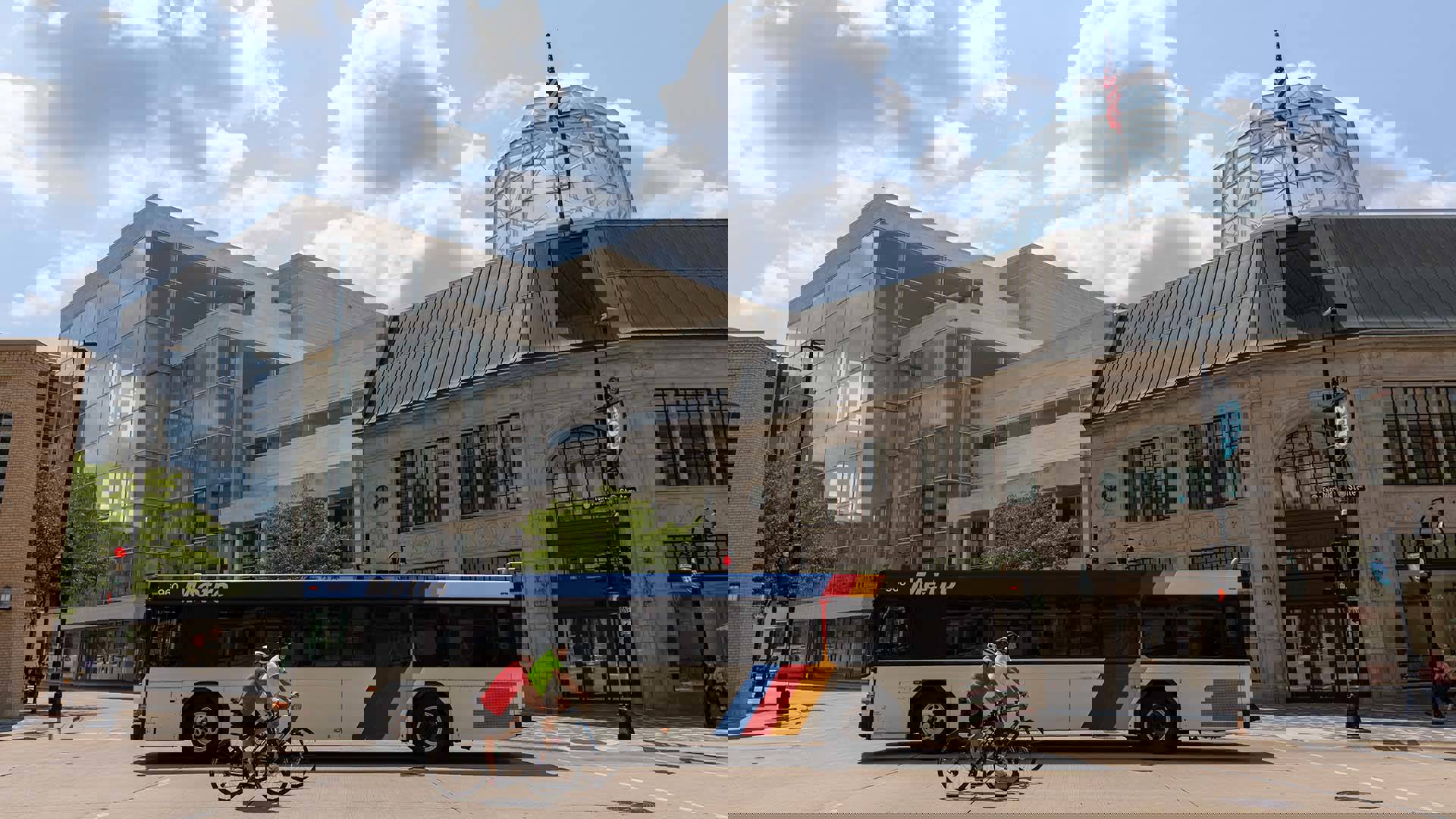 A Madison Metro bus and two bikers crossing Fairchild St on State St in front of Overture Center on a bright summer day.