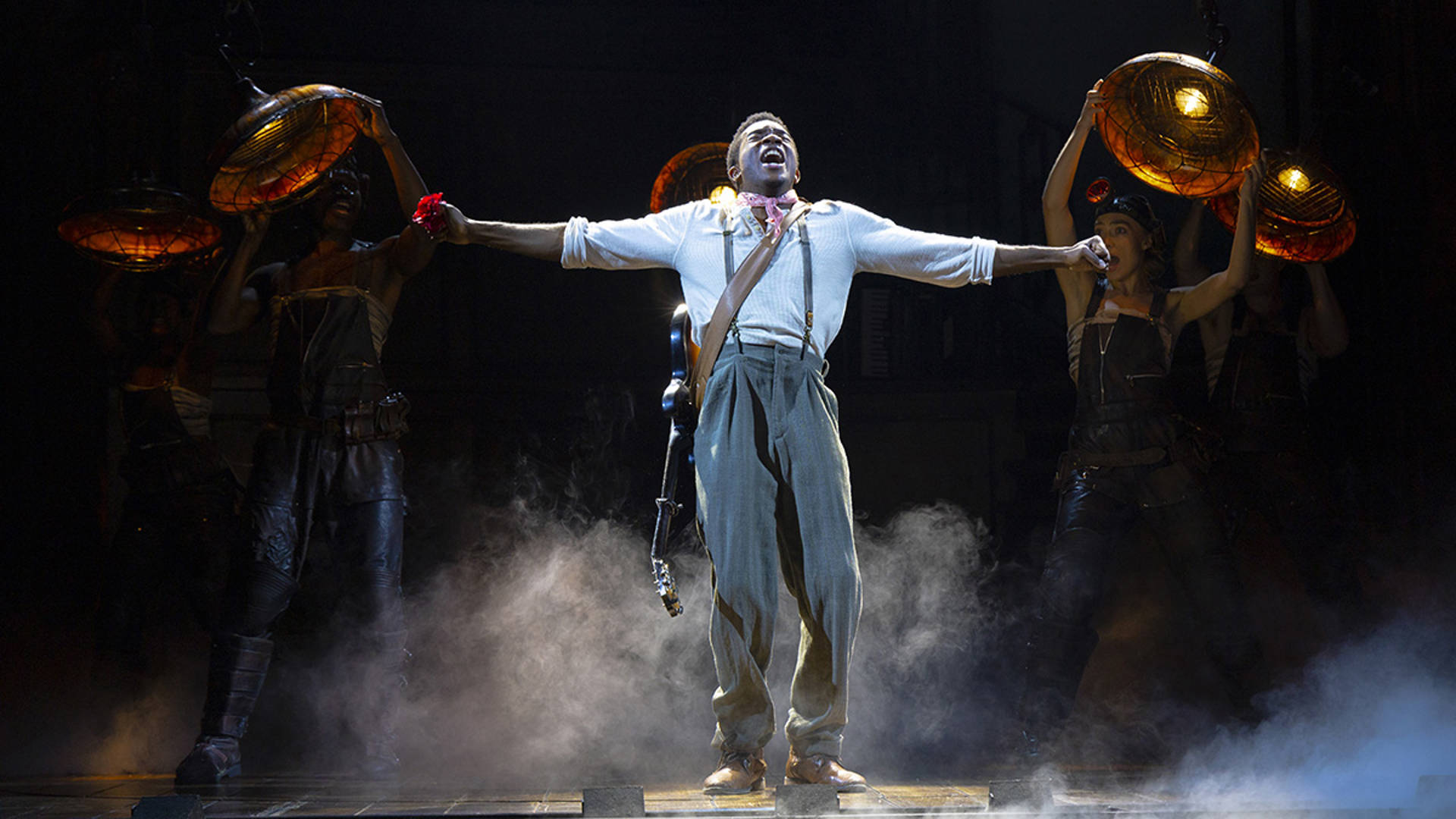 An African American man in a white shirt and suspenders sings with his arms outstretched. He has a guitar slung around is shoulders.