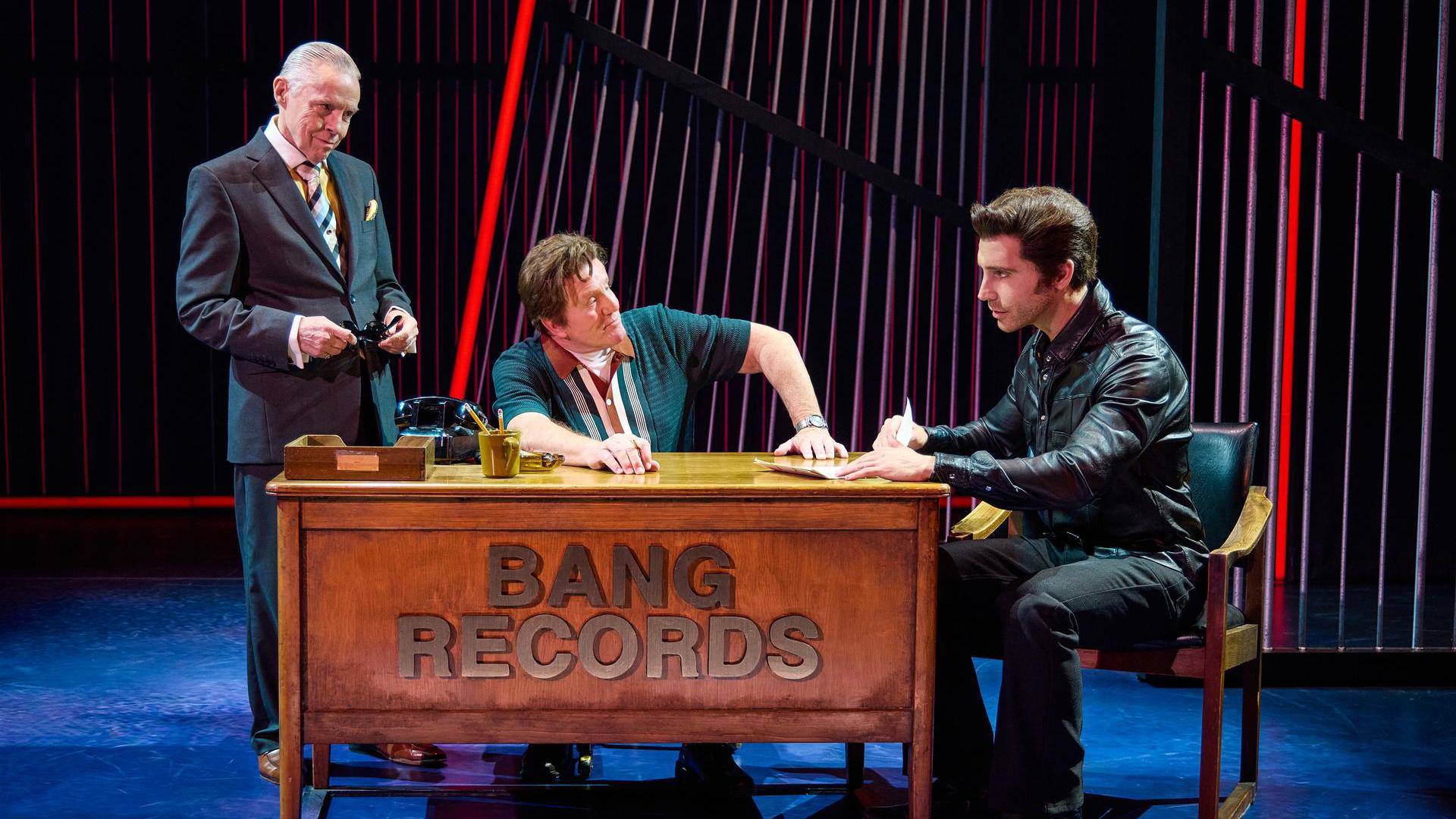 A man in a dark outfit with light complexion and greased hair signing a contract at a desk as two other men look on. The desk says, "Bang Records"