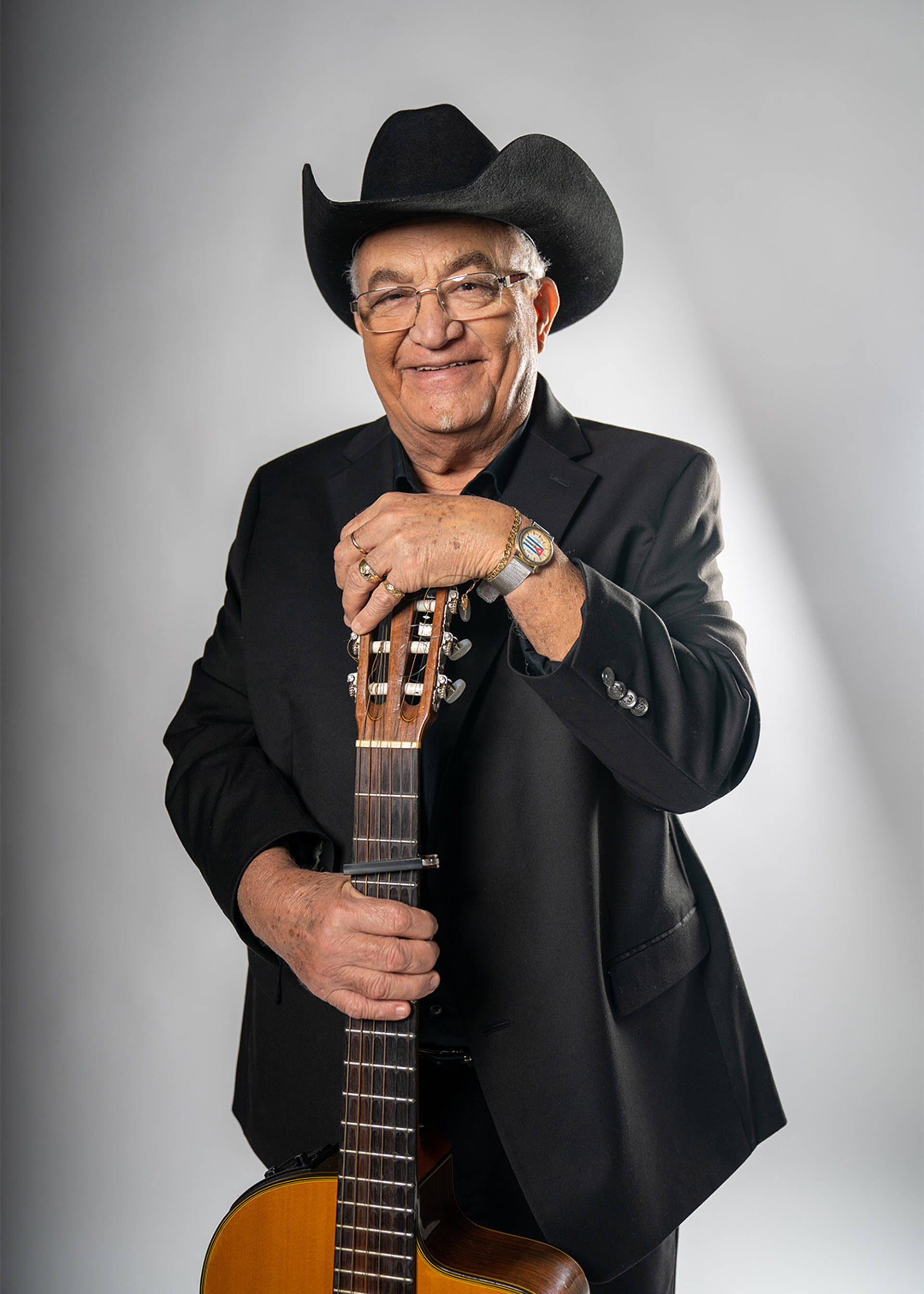 Headshot of Eliades against a white background. An older Cuban man with medium skin tone and short white hair. He is wearing glasses, a black cowboy hat and an all black suit while smiling and holding a guitar.