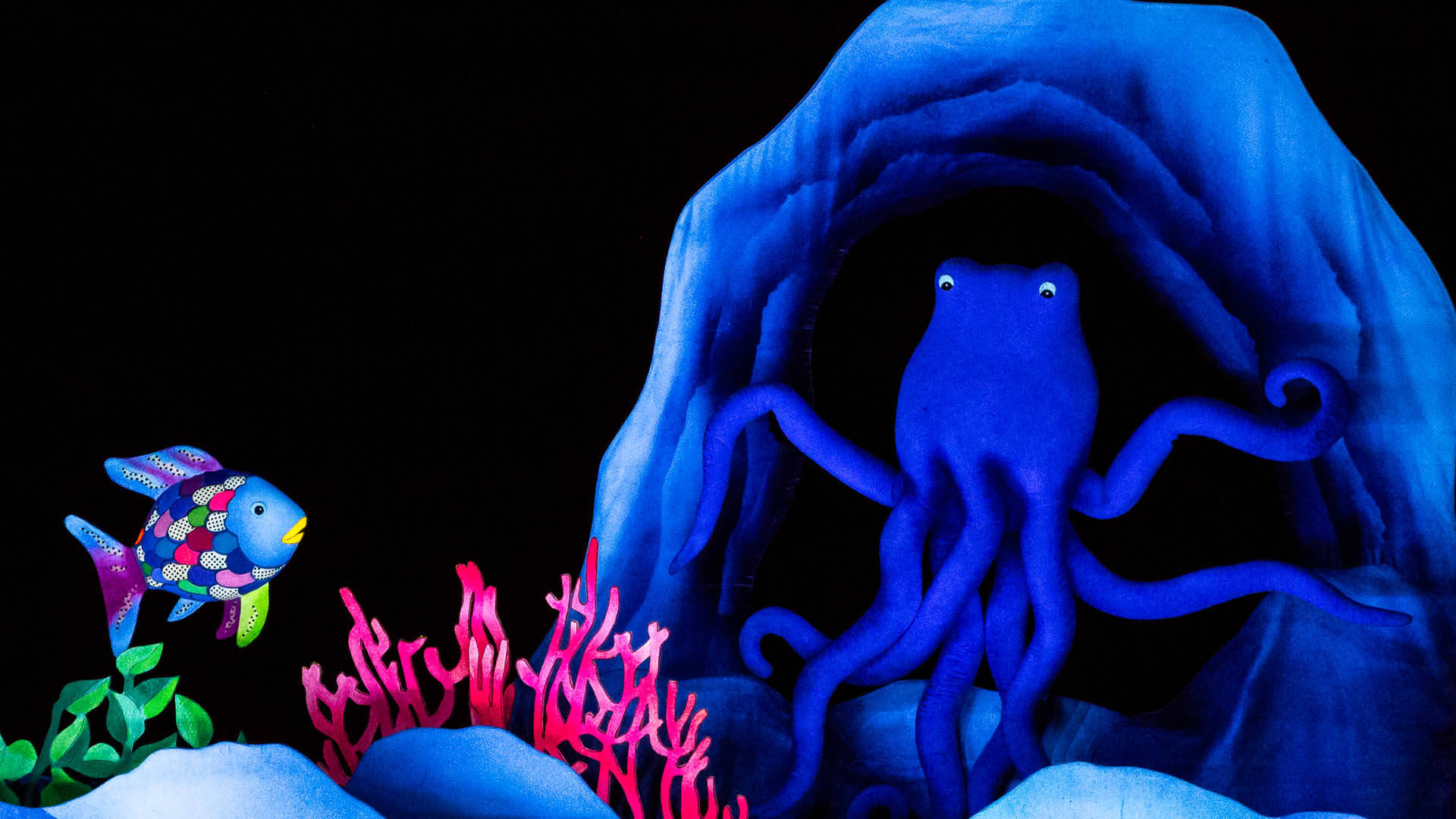 Puppets of a fish with rainbow scales and a large, blue octopus. The octopus is in the entrance to a cave. The rainbow fish is to the left of the octopus, above some pink coral and green seaweed.