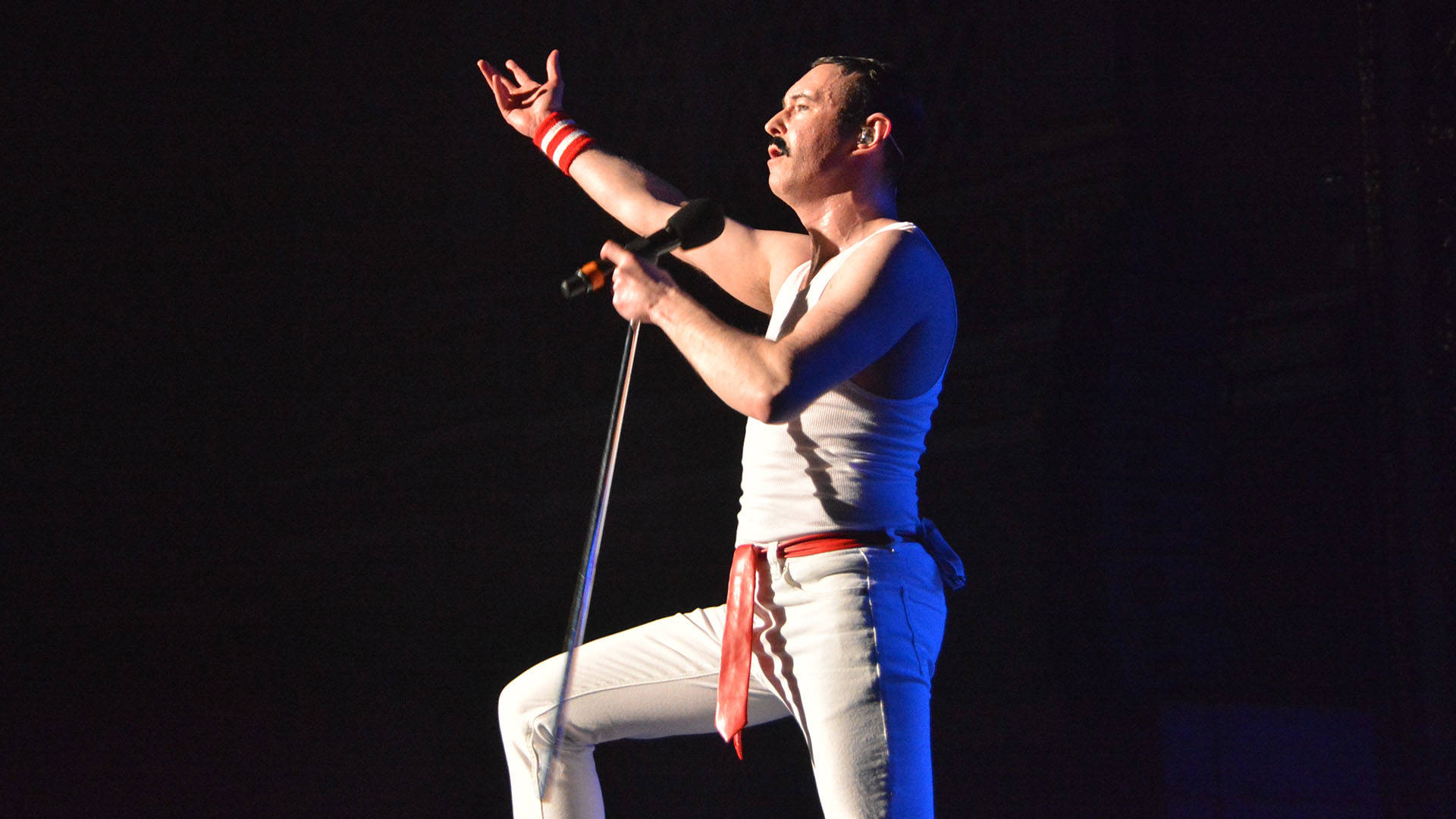 A caucasian man with short black hair and a moustache singing into a microphone with his right hand outstretched. He is wearing all white with a red belt.