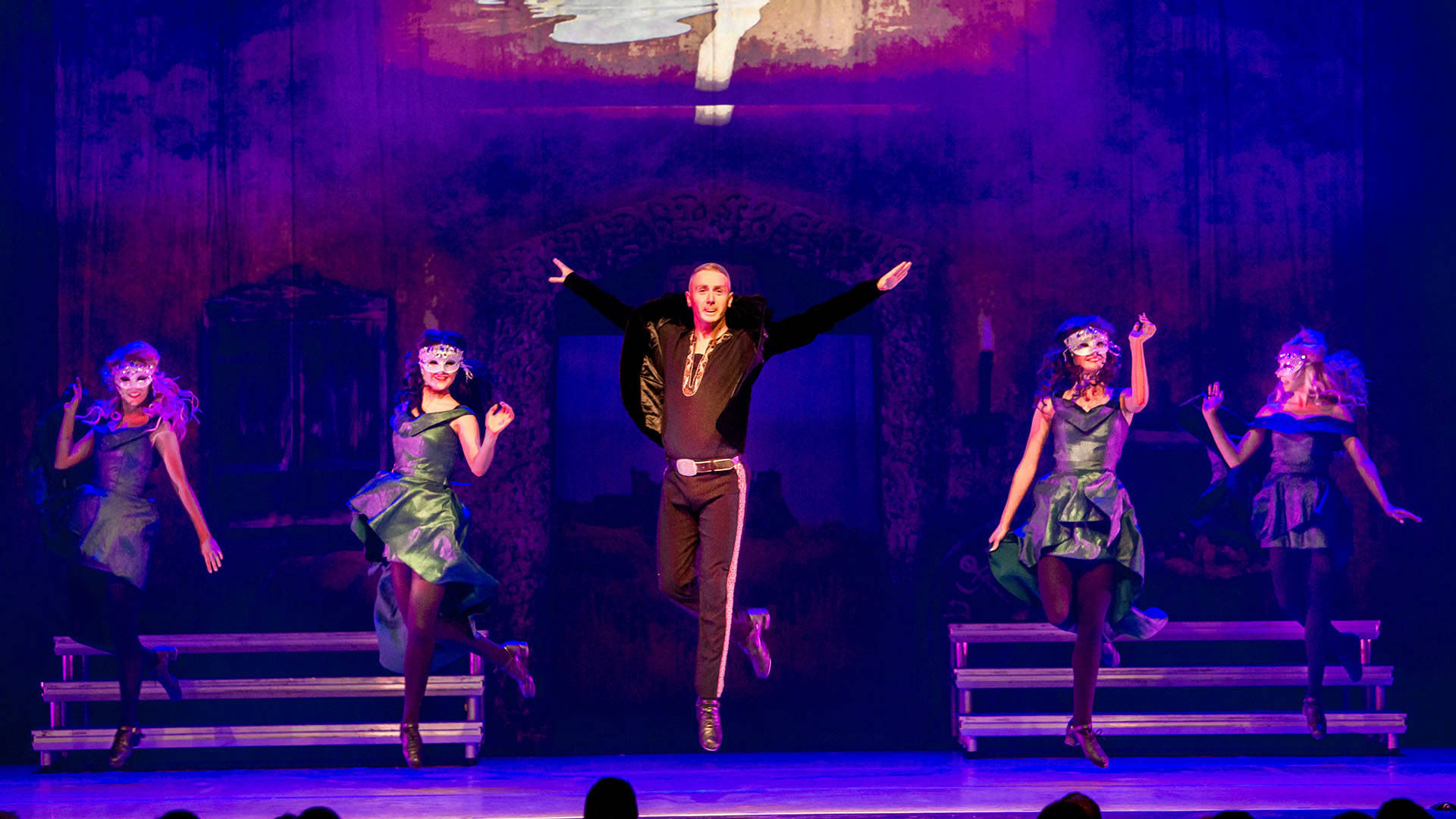 A group of Irish dancers in black and green outfits performing on stage. The center dancer is leaping into the air.