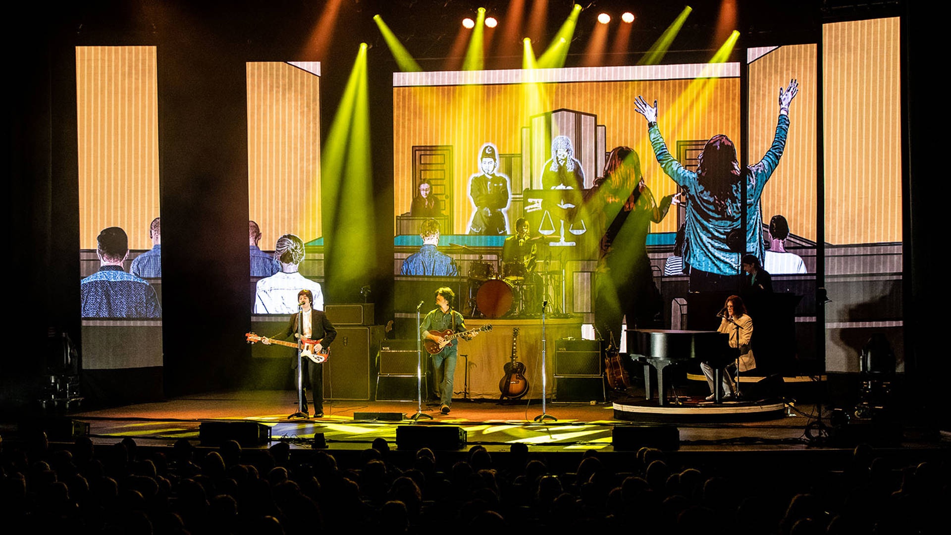 Four Beatles impersonators performing in front of a full house with guitars, a piano and a drum set. A cartoon court room scene is projected behind them.