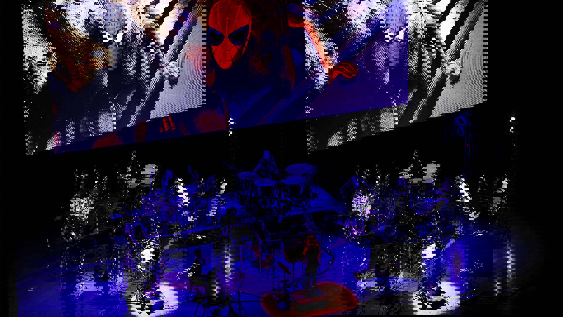 View from above of an orchestra performing on stage with a large screen above them. A scene from Spiderman: Into the Spider-verse is playing