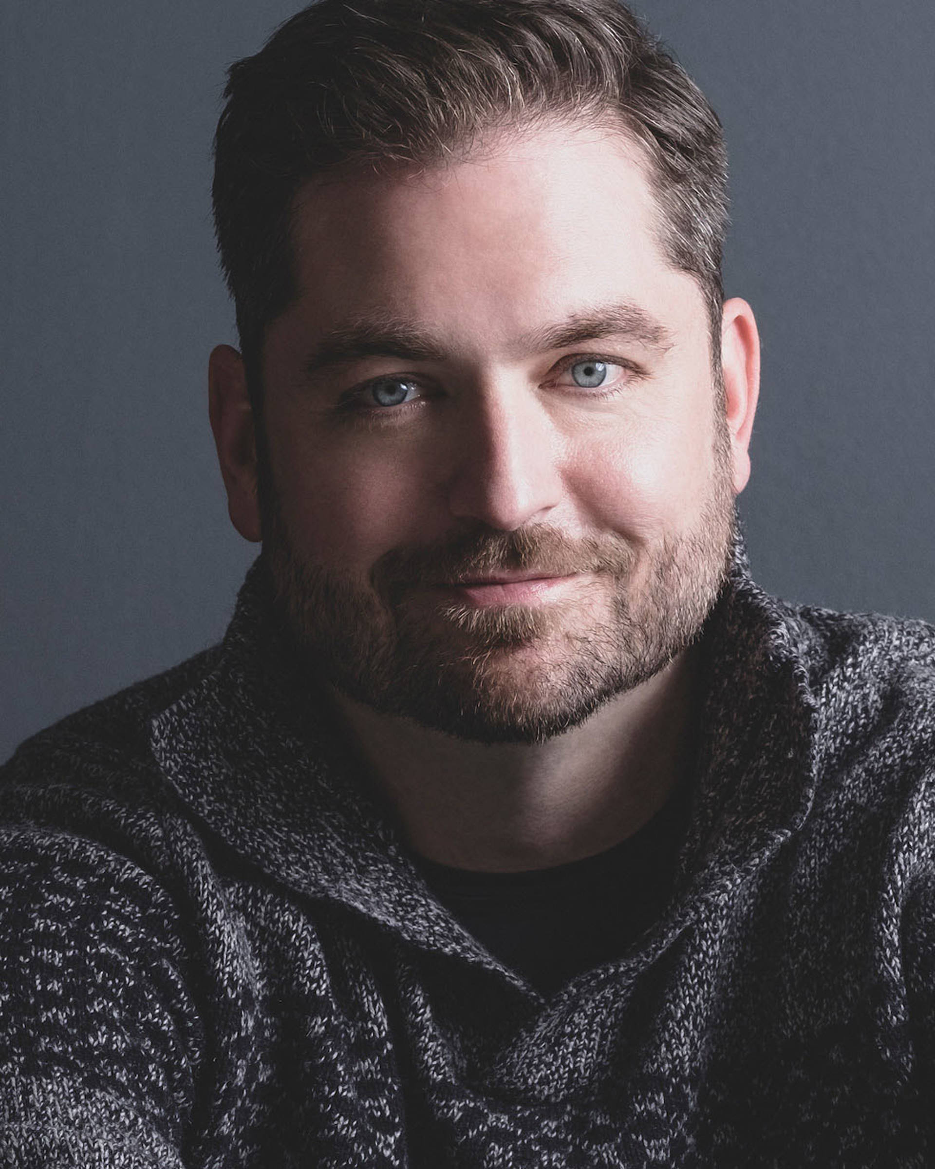 Headshot of a caucasian man with short dark hair, blue eyes, and a beard. He is wearing a sweater.