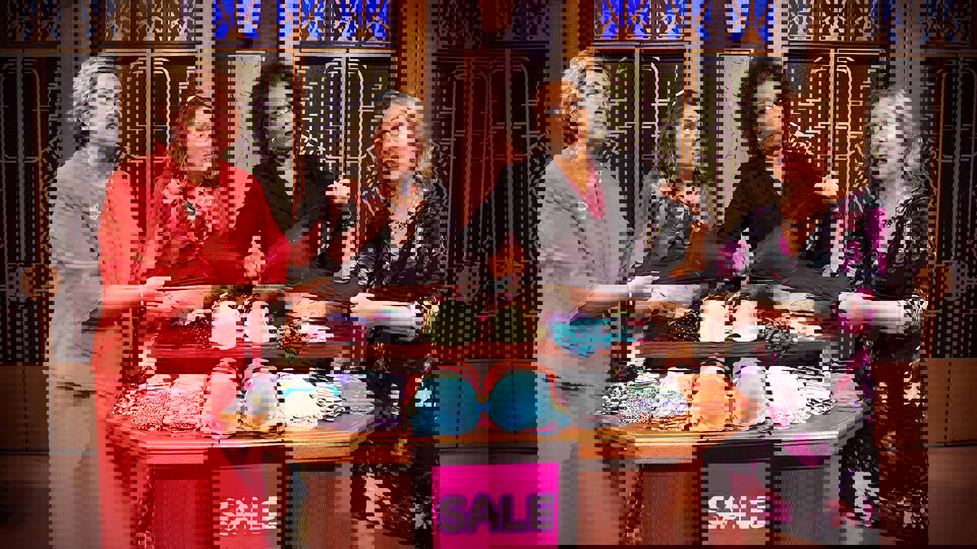 Four women make funny faces at eachother as they fight over a black bra.