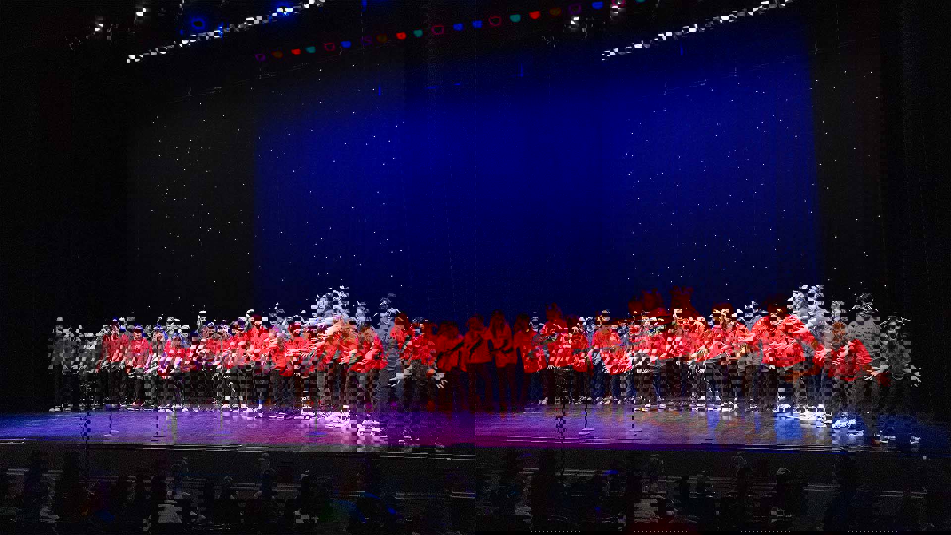 Young students in matching red shirts and dark pants performing on a dark stage. 