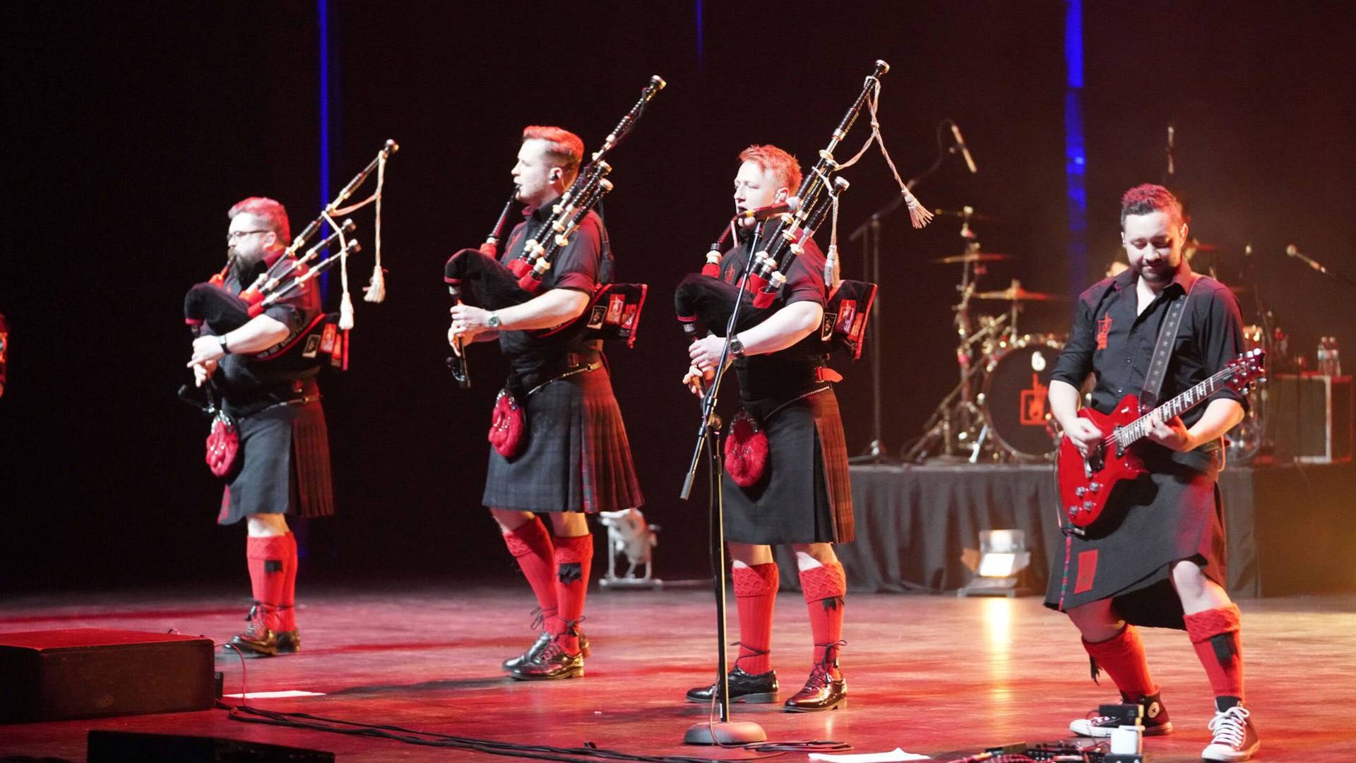 Four musicians on stage, three playing bagpipes and one with an electric guitar, all wearing matching tartan kilts.