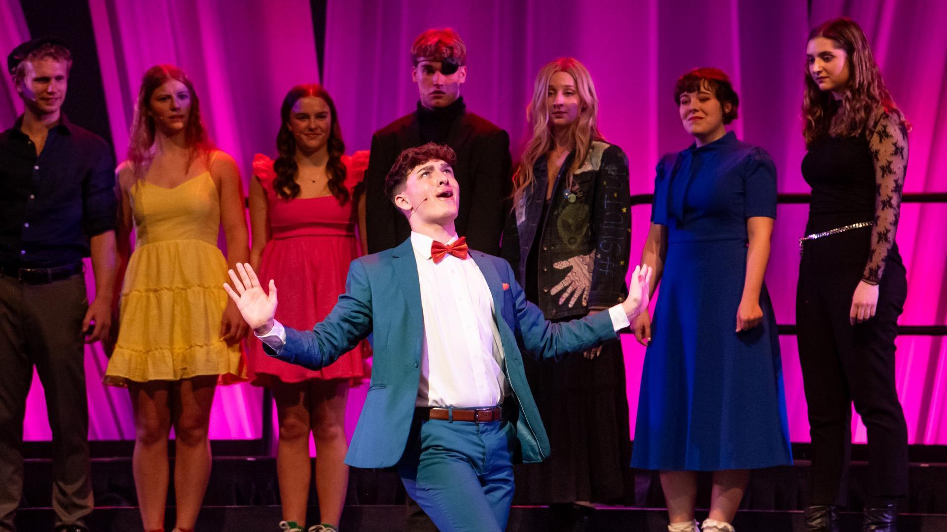 A high school aged performer in a blue suit and red bowtie performing on a theater stage with his hands held out. Other well dressed high schoolers are standing behind him