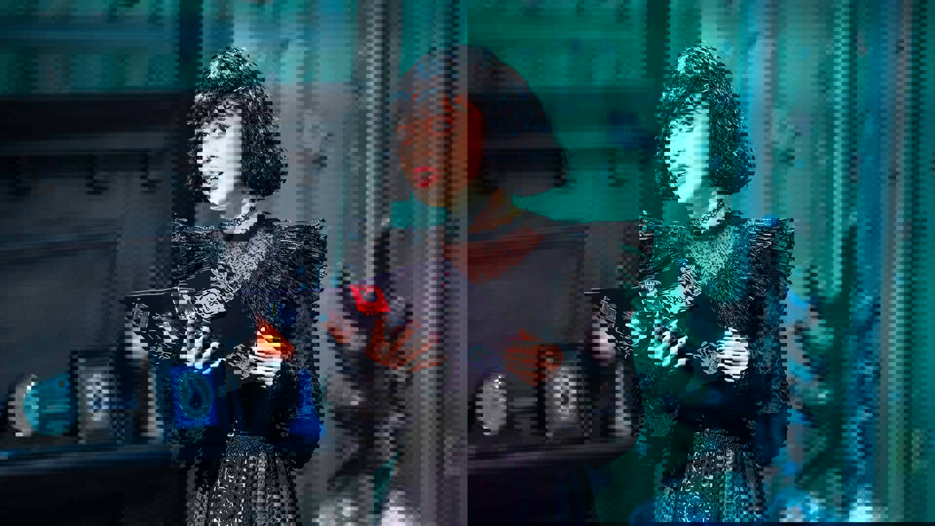 A Columbian-American young woman performing on stage. She has a black hair with bangs and is wearing a black frilly dress and holding open a black binder with stickers.