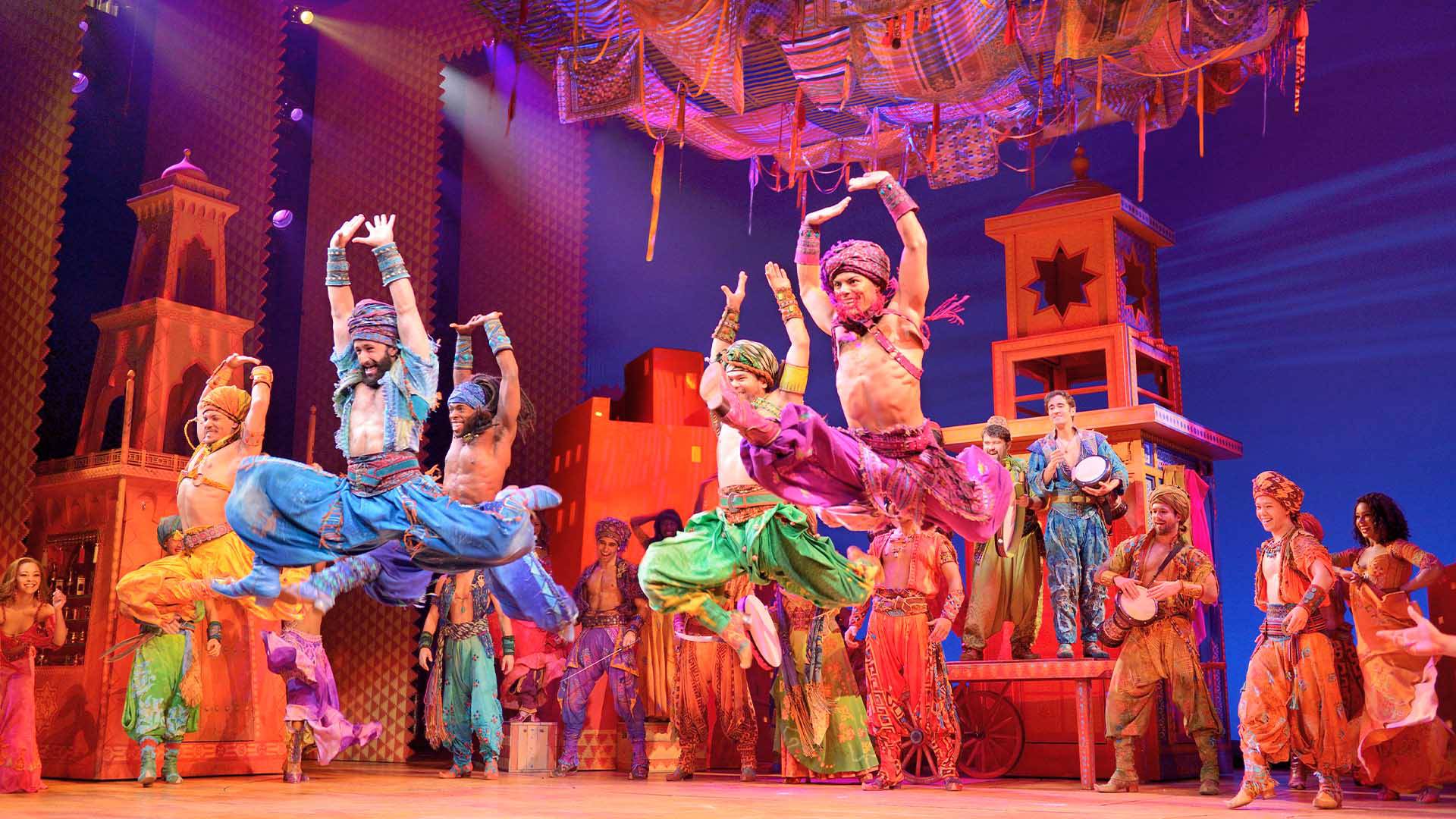 Men in Middle Eastern style clothing leaping in the air. Many other actors watch them in the background. They are in a bazaar at night time.