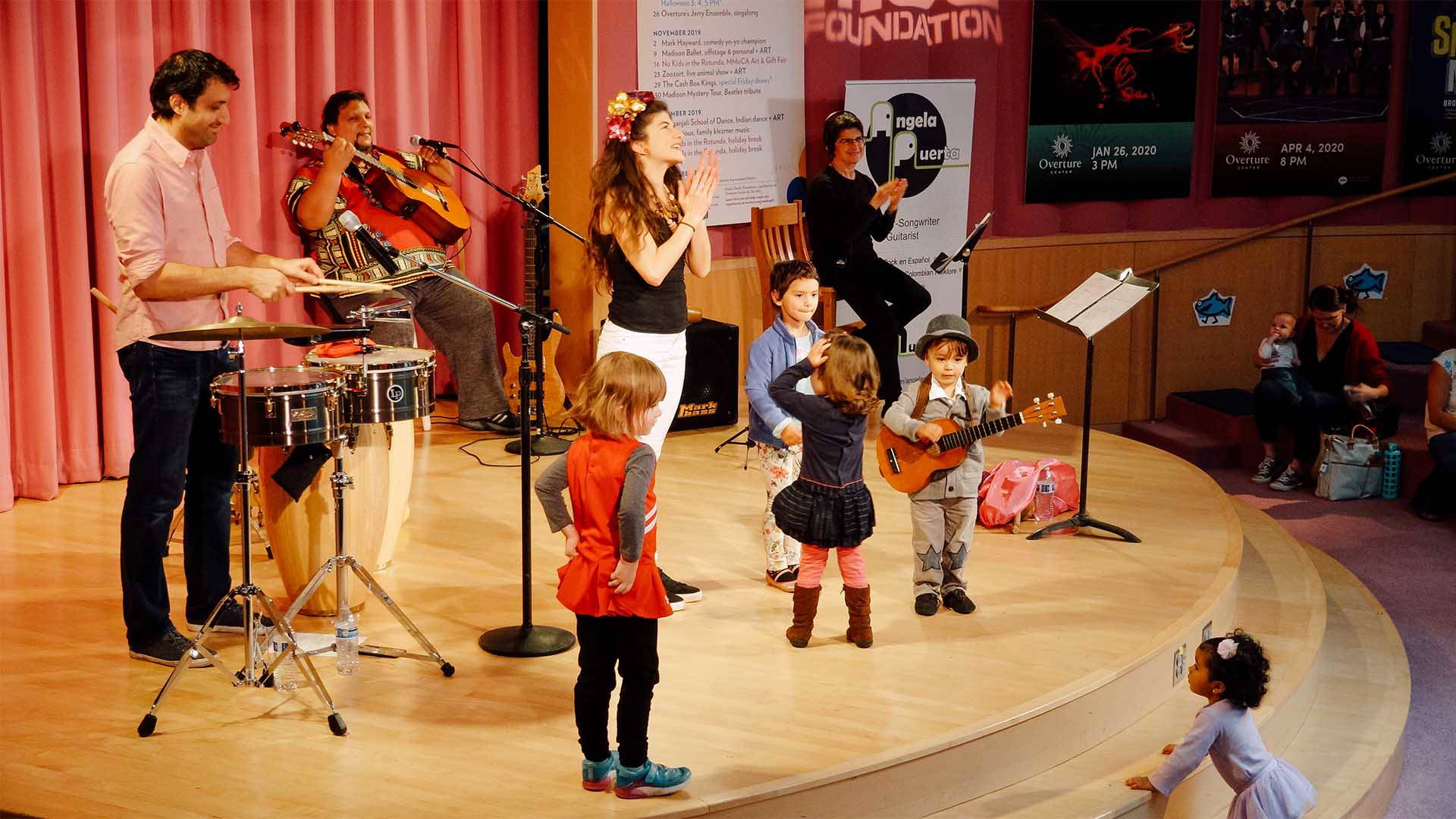 Several musicians on a small stage. One is playing timbales, another guitar. Several small children join them on stage.