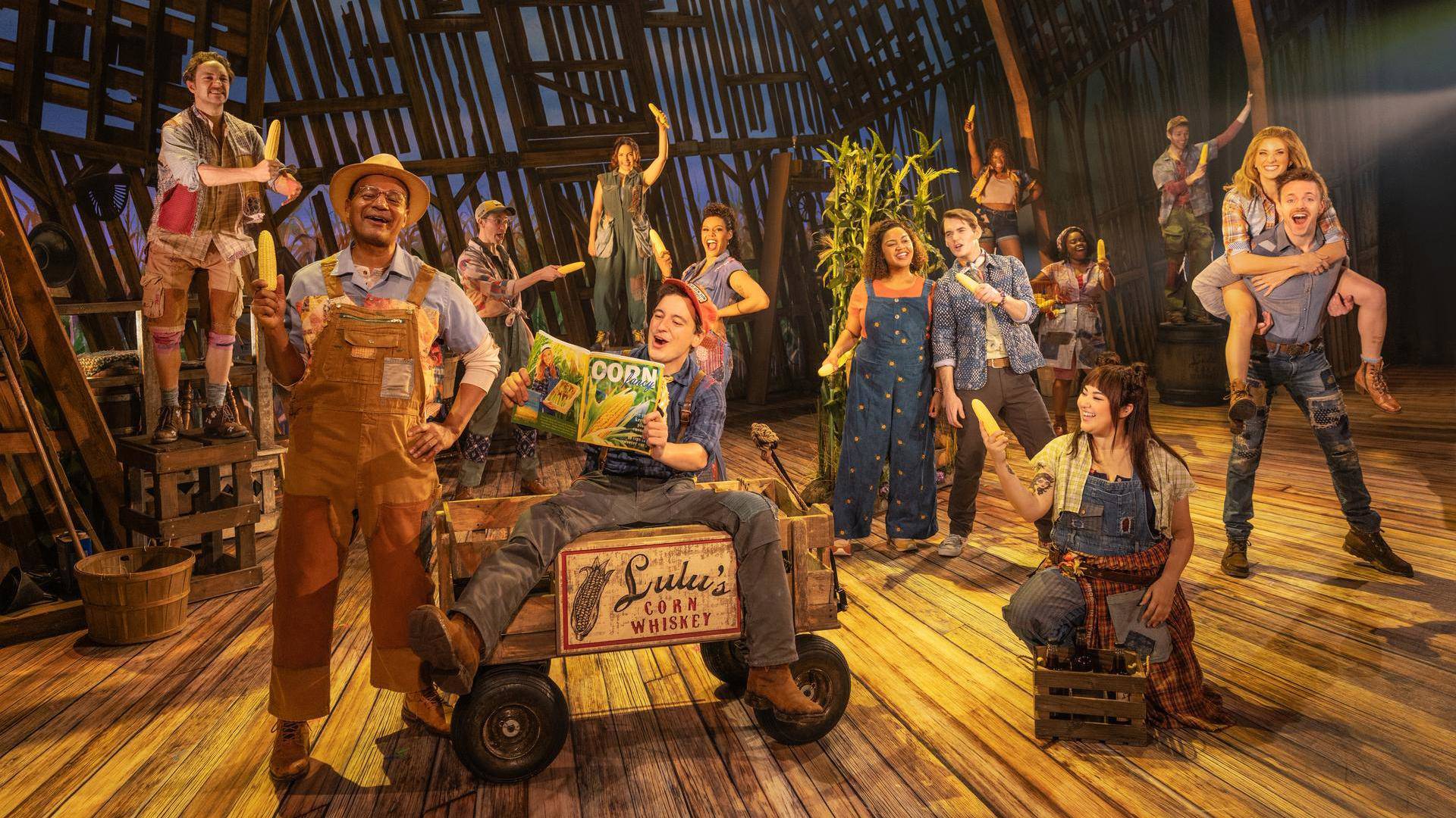 A group of people in various poses singing and wearing farmwork outfits on a wooden floor. Many are holding heads of corn.