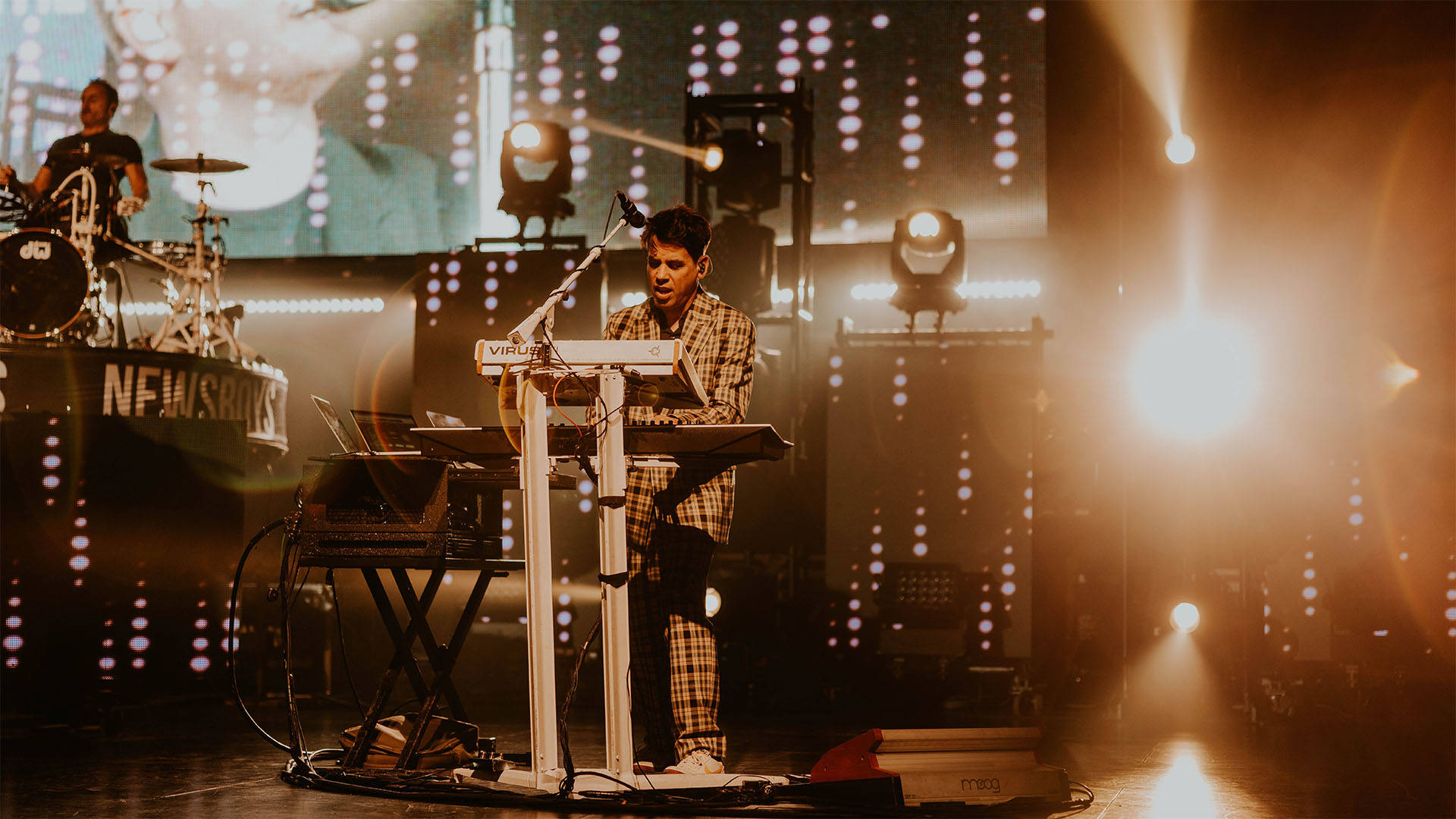 A man with light complexion, short dark hair and a plaid suit playing mounted keyboards on a stage.