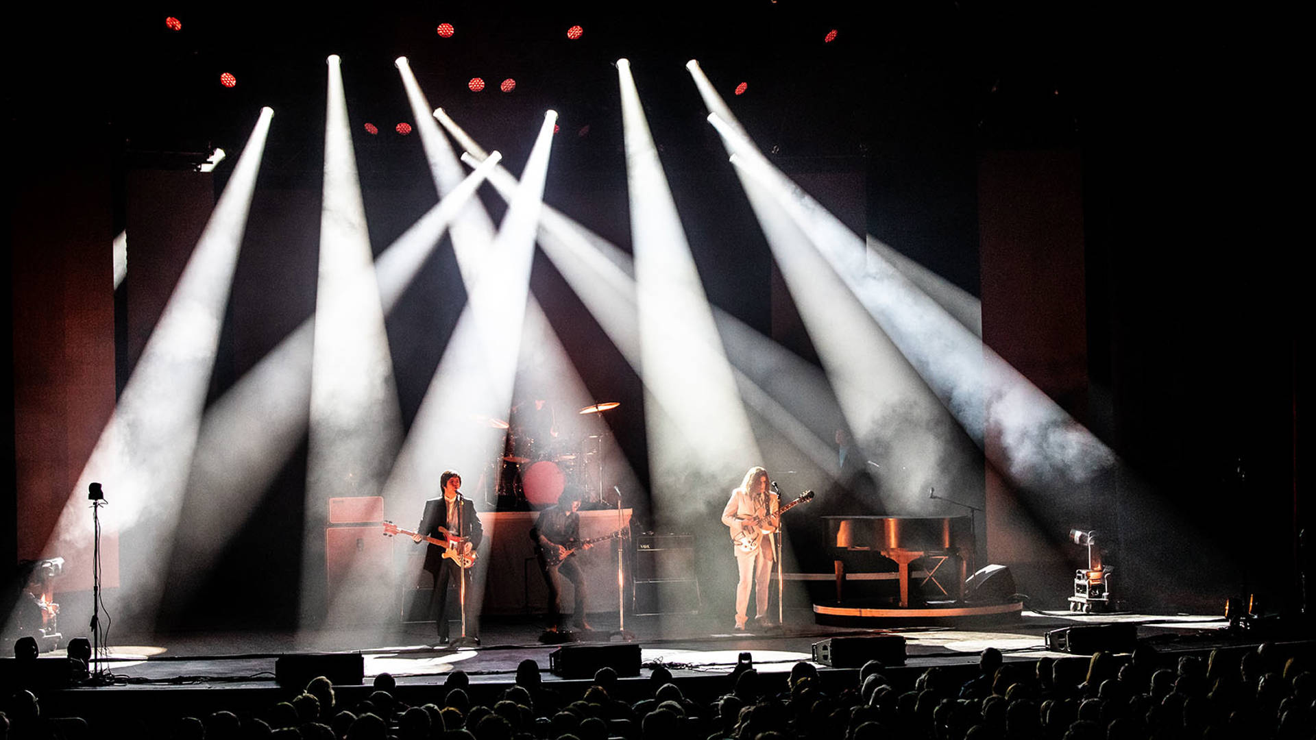 Four Beatles impersonators performing on a stage with streaking spotlights aroud them, wearing stylish 1960s street clothes.
