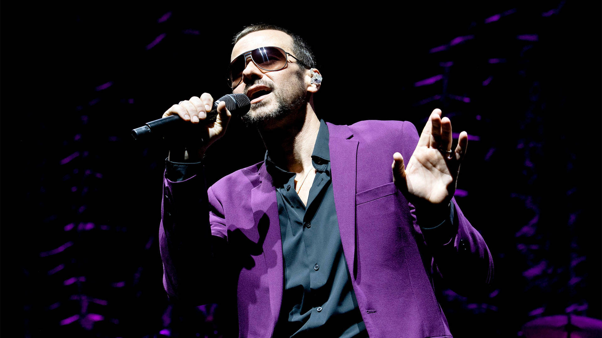 A white man with short brown hair, sunglasses, and a purple jacket singing into a microphone.